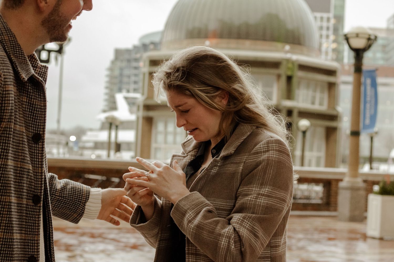 Emotional woman admiring engagement ring after proposal at rainy waterfront location.