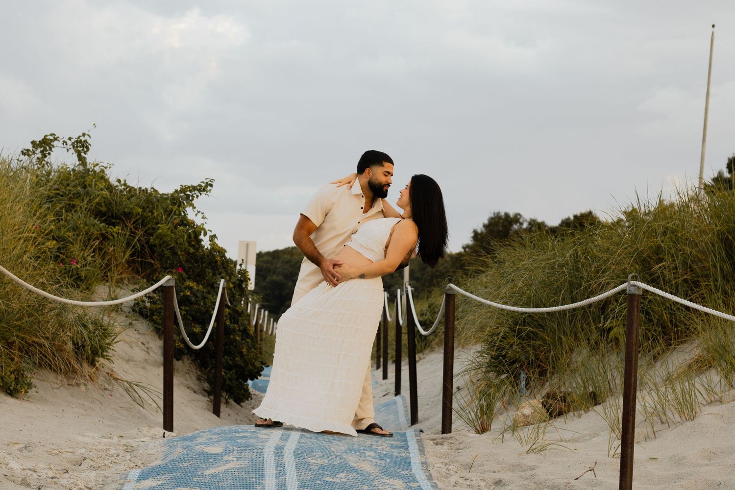 Romantic couple almost kissing on a sandy beach pathway lined with rope railings and dune grass.