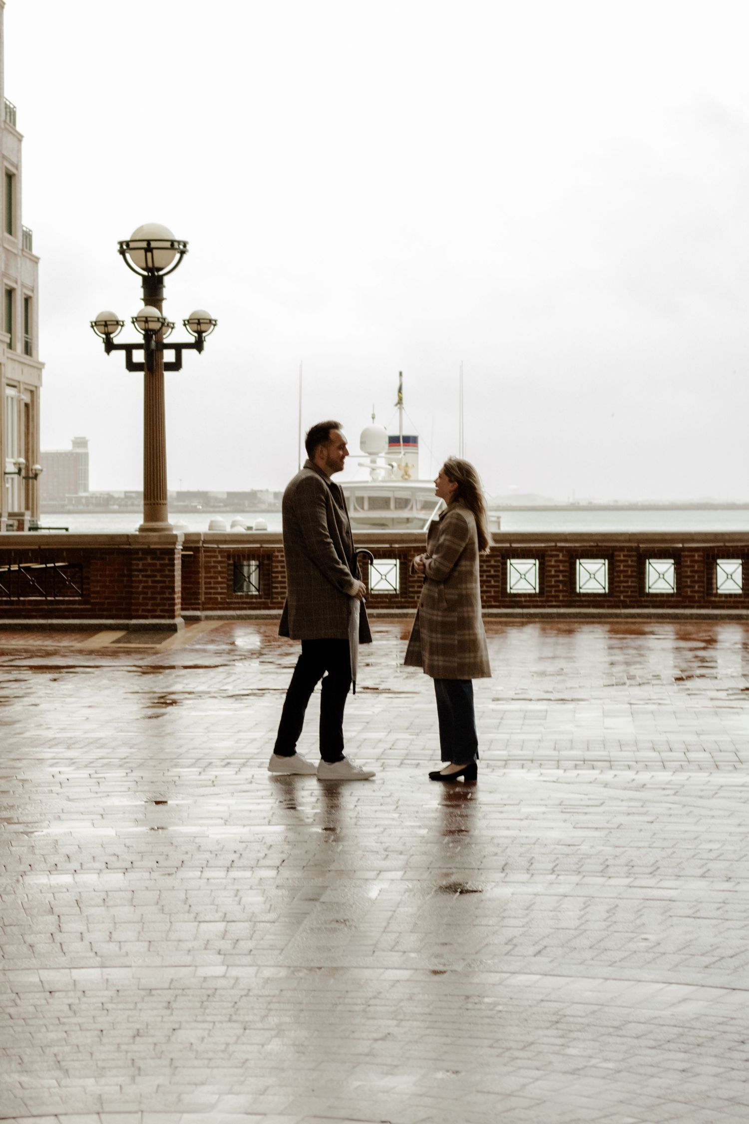 Couple standing face-to-face on wet cobblestone waterfront plaza on a cloudy, overcast day.