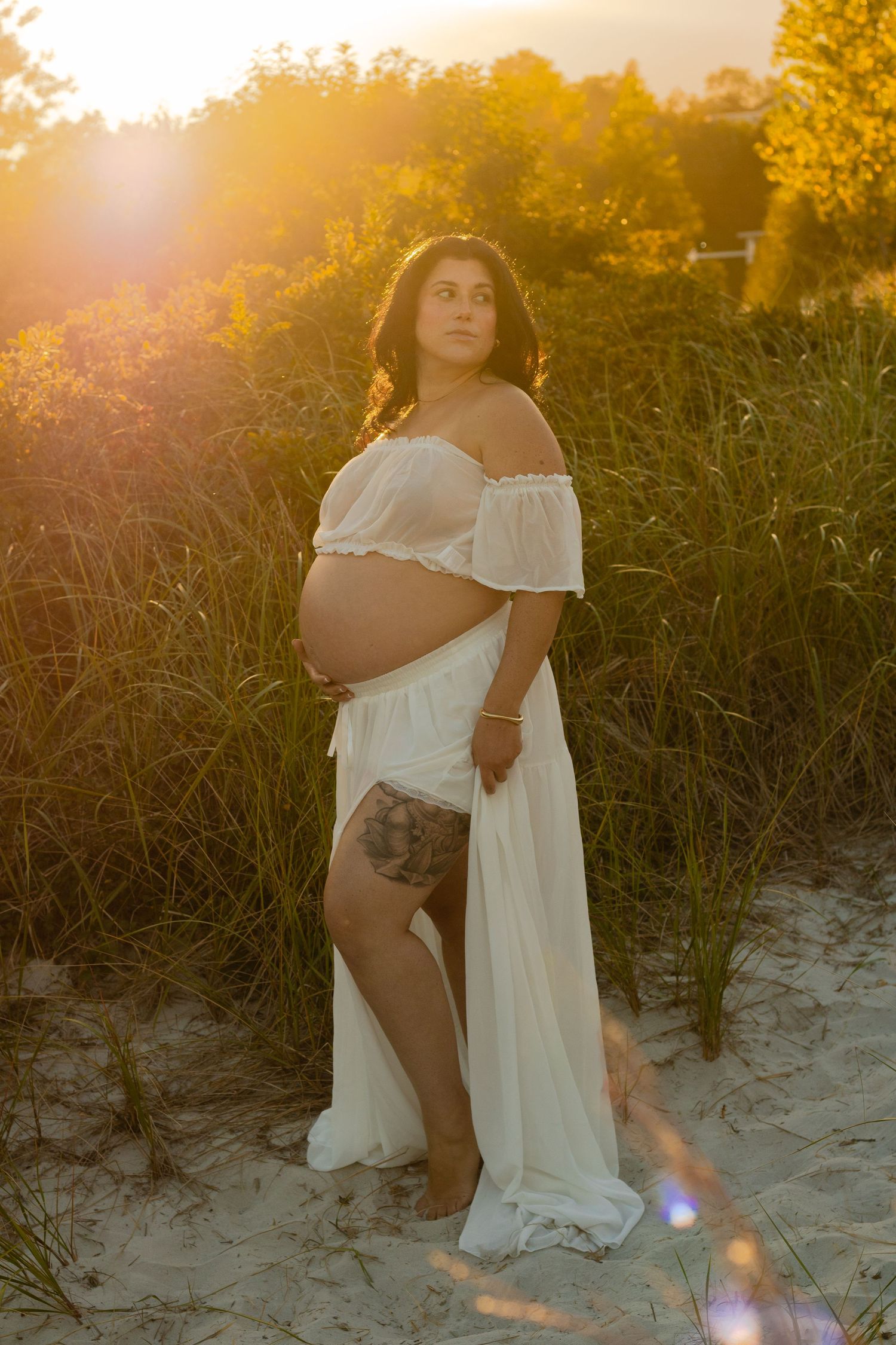 Pregnant woman in white off-shoulder gown poses outdoors at golden sunset, showing baby bump amid tall grass.