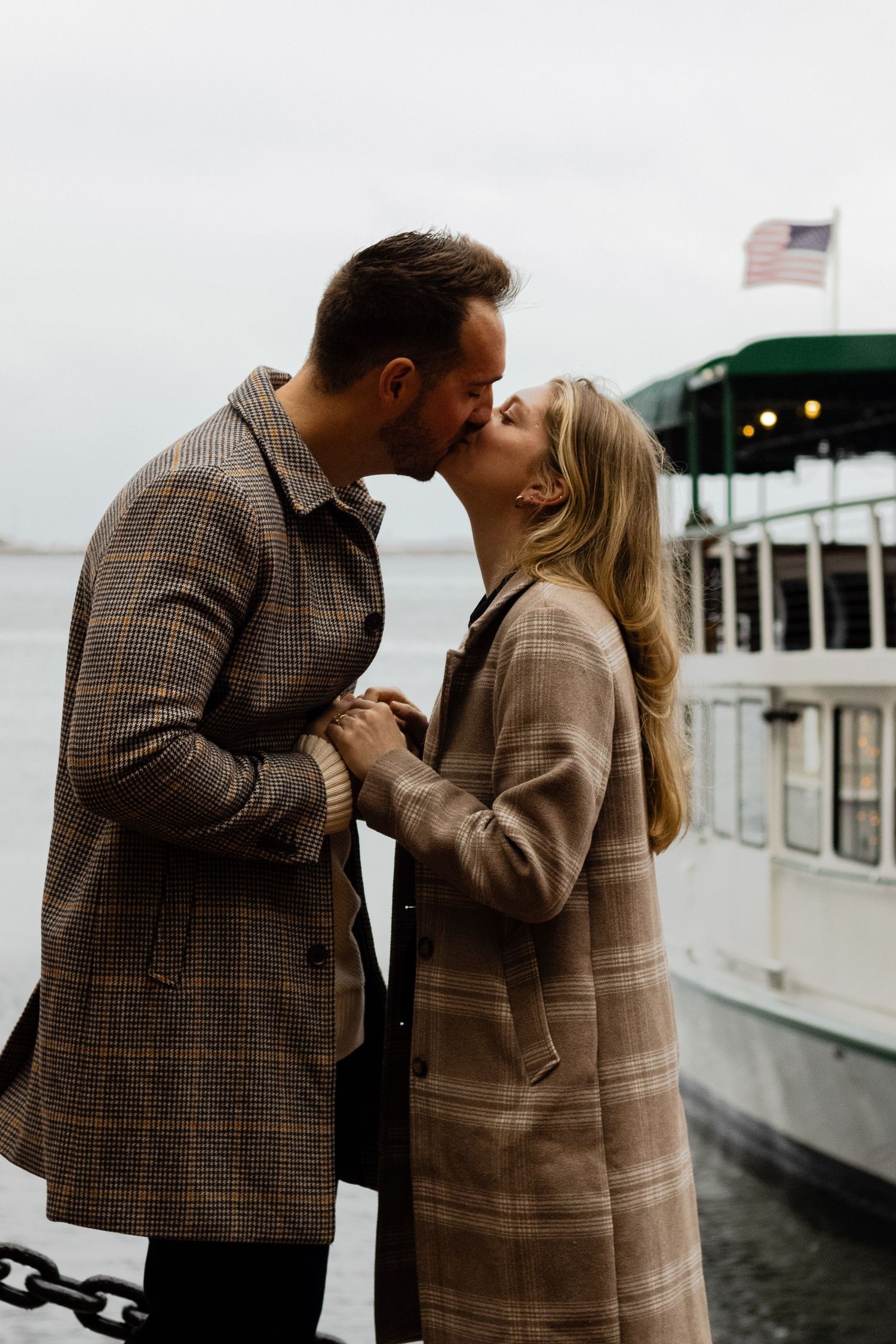 A romantic couple shares a kiss near a docked boat on a cloudy waterfront day.