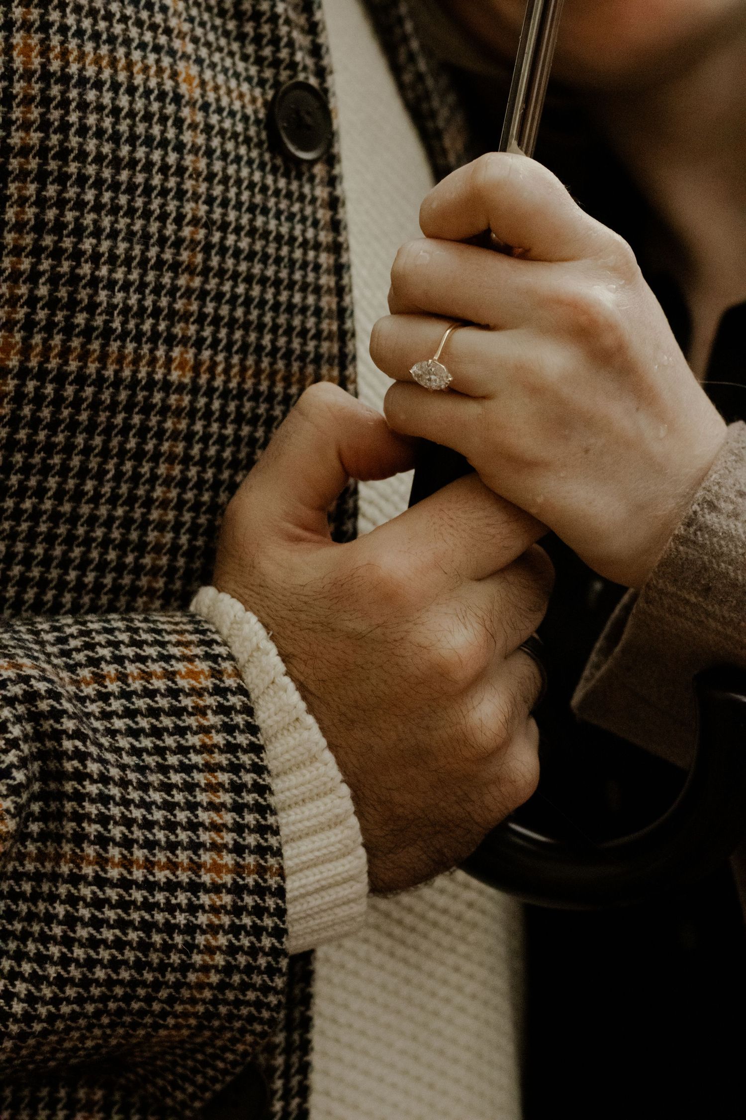 Close-up of an engagement ring on a woman's hand as a couple holds hands near a plaid jacket.