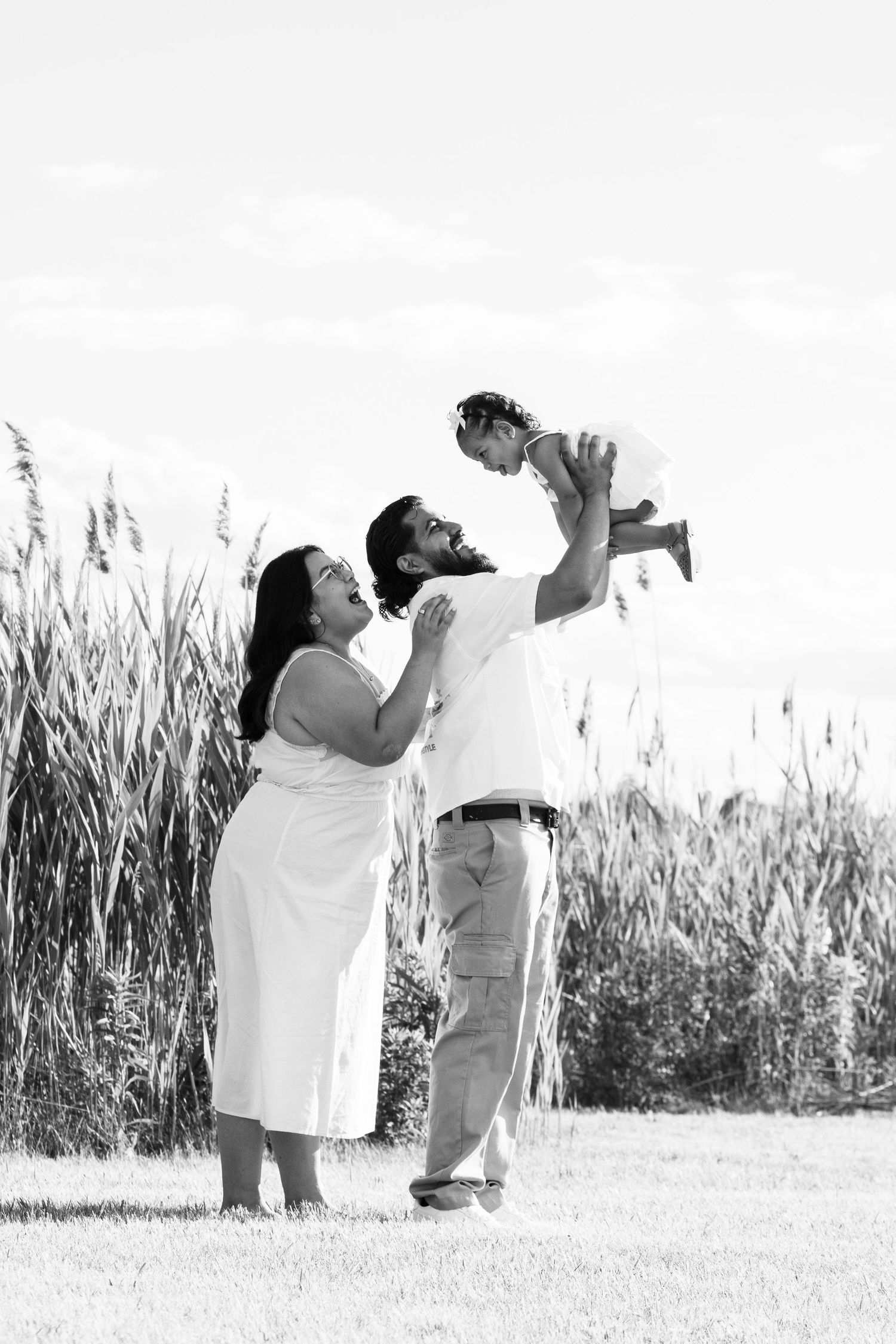 Black and white photo of parents joyfully lifting their baby in the air in a grassy field.