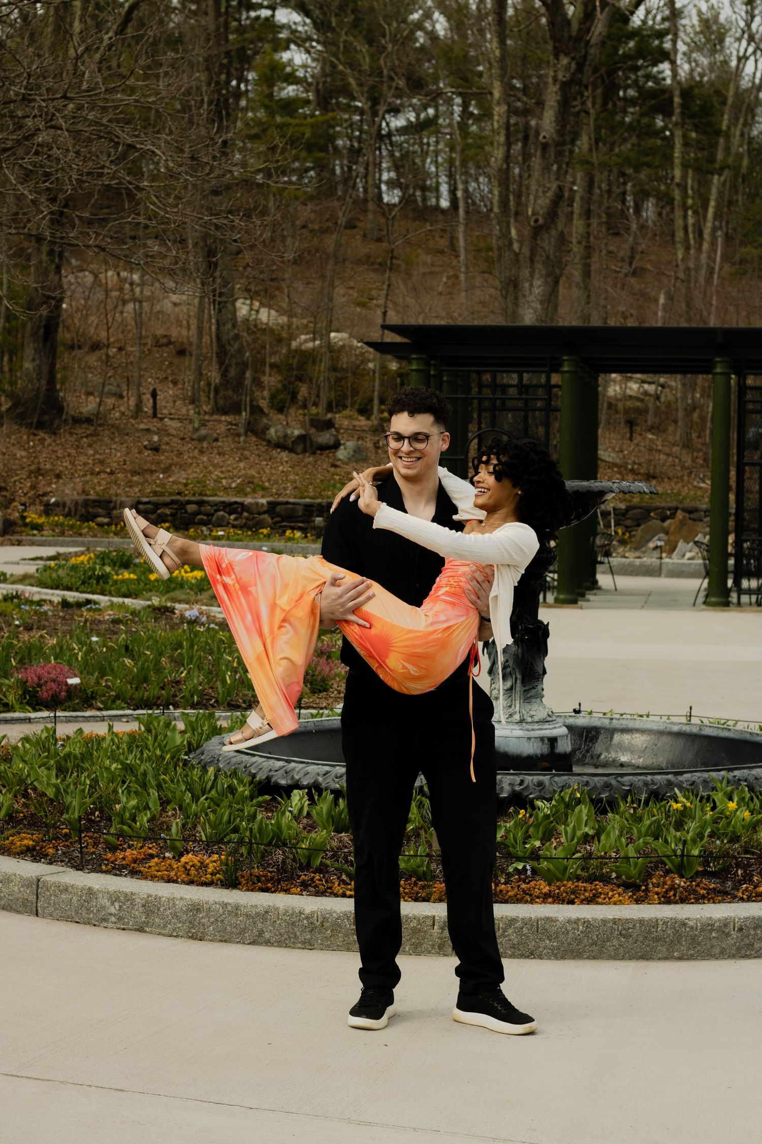 A man in black carries a woman in a colorful dress by a garden fountain, both smiling outdoors.