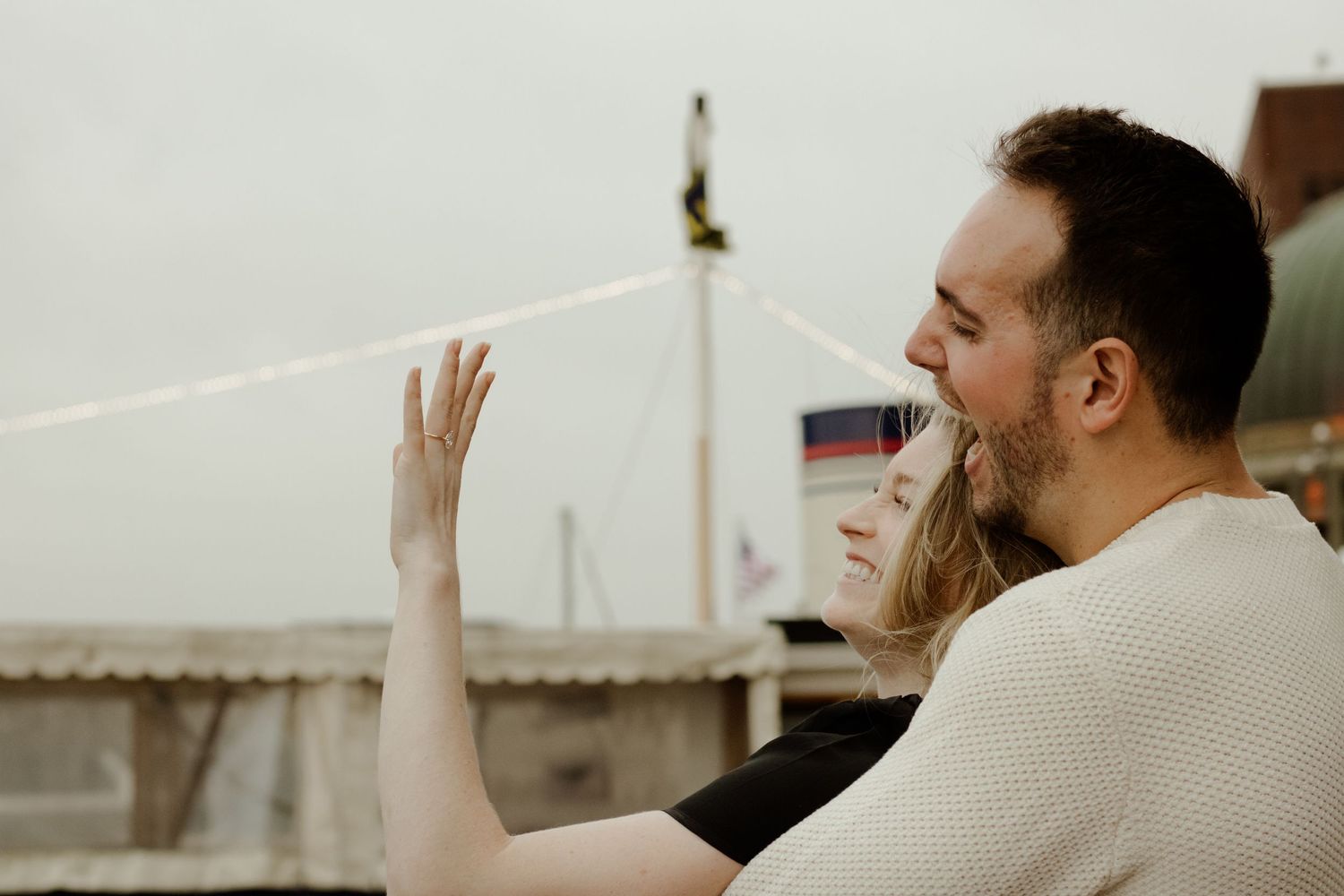 Couple embracing on a rooftop, woman waving her hand while leaning into mans arms on an overcast day.