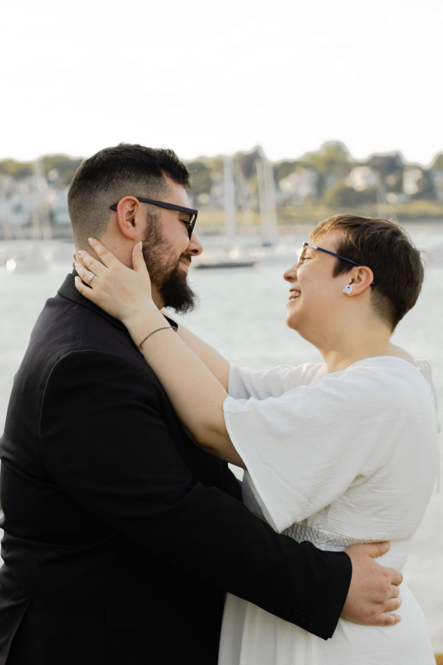 Smiling couple embracing near a marina, woman in white dress touching mans bearded face.