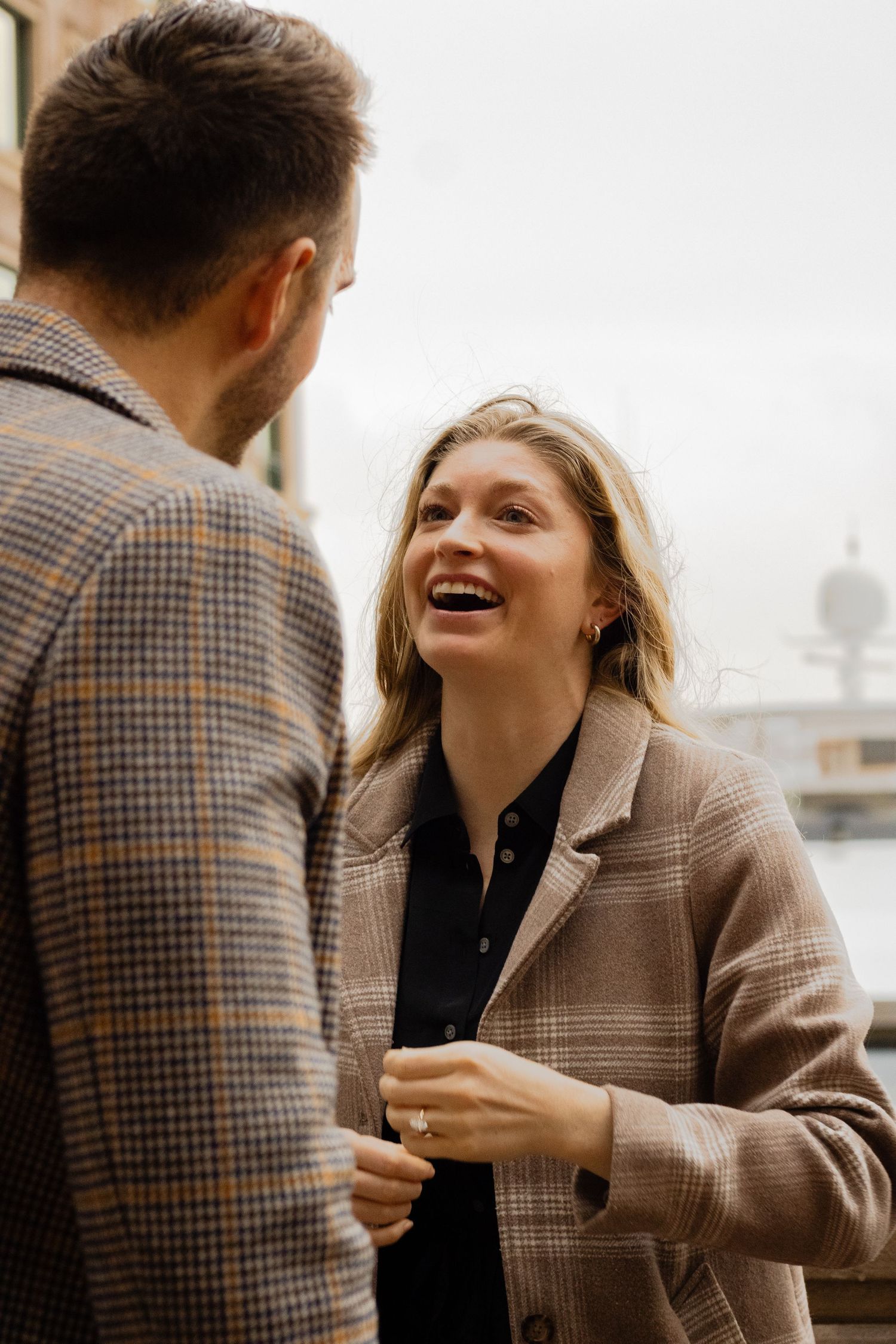 A laughing blonde woman in a plaid coat faces a man outdoors near a waterfront.