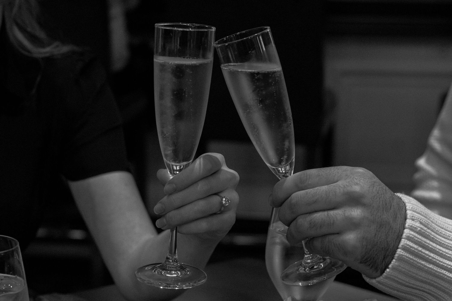 Black and white close-up of a couple toasting with champagne flutes, engagement ring visible.