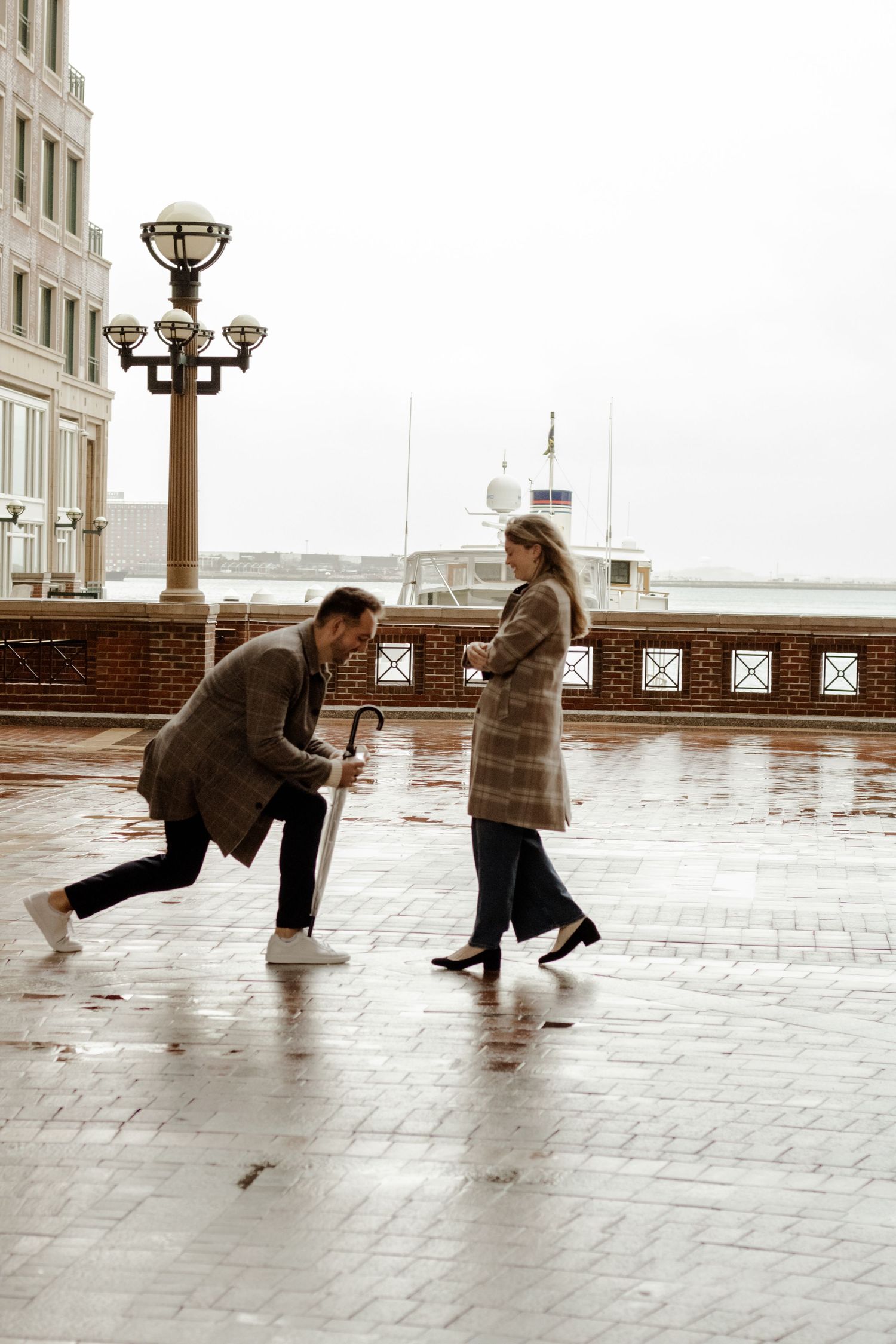Man playfully chasing woman on a rainy waterfront promenade, both wearing coats on an overcast day.