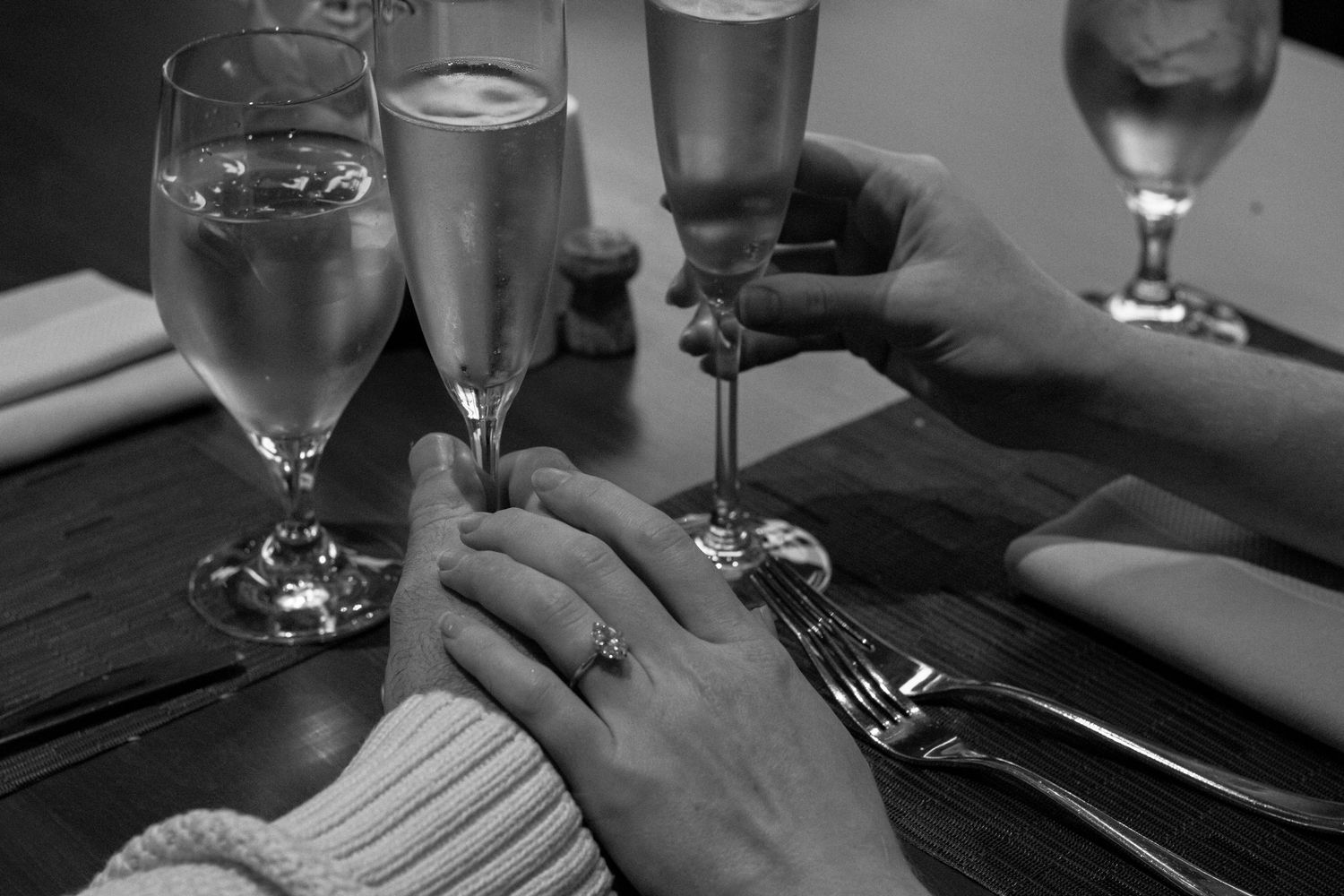 Black and white close-up of engaged couple holding hands and toasting with champagne at a restaurant.