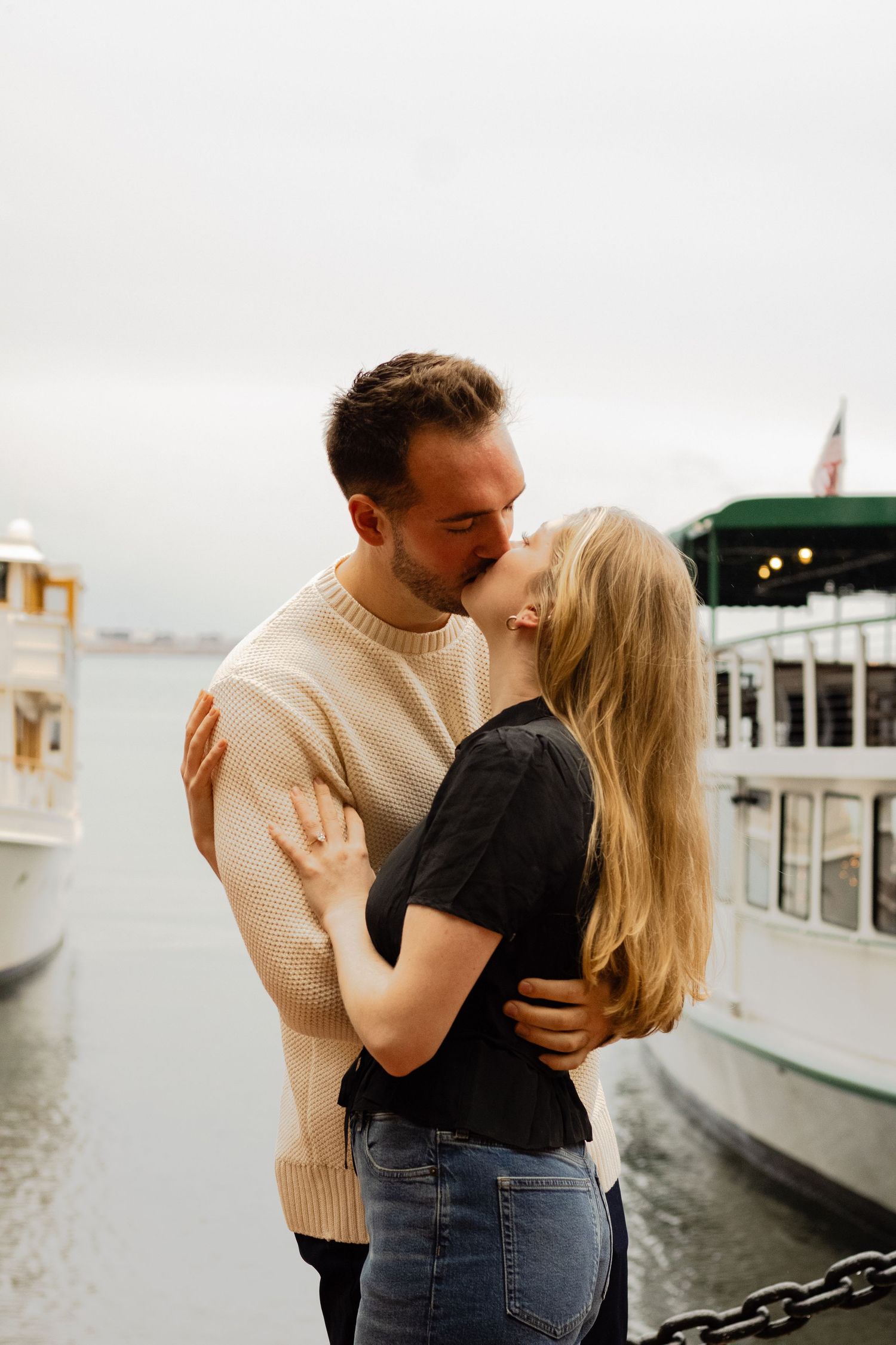 A couple shares a romantic kiss while embracing near docked boats on a cloudy waterfront.