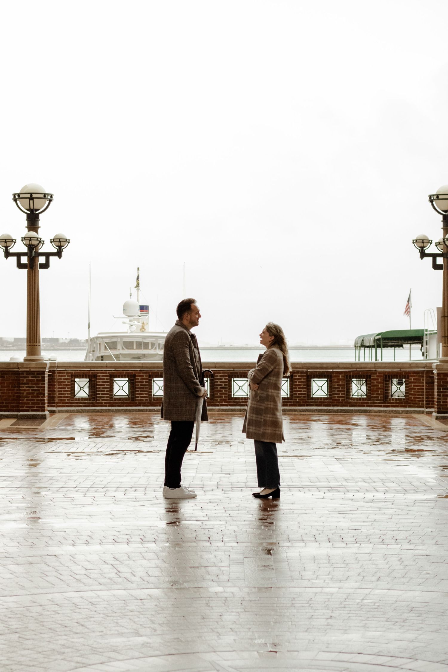 Couple facing each other on a rain-soaked waterfront promenade with lamp posts and a ship in background.