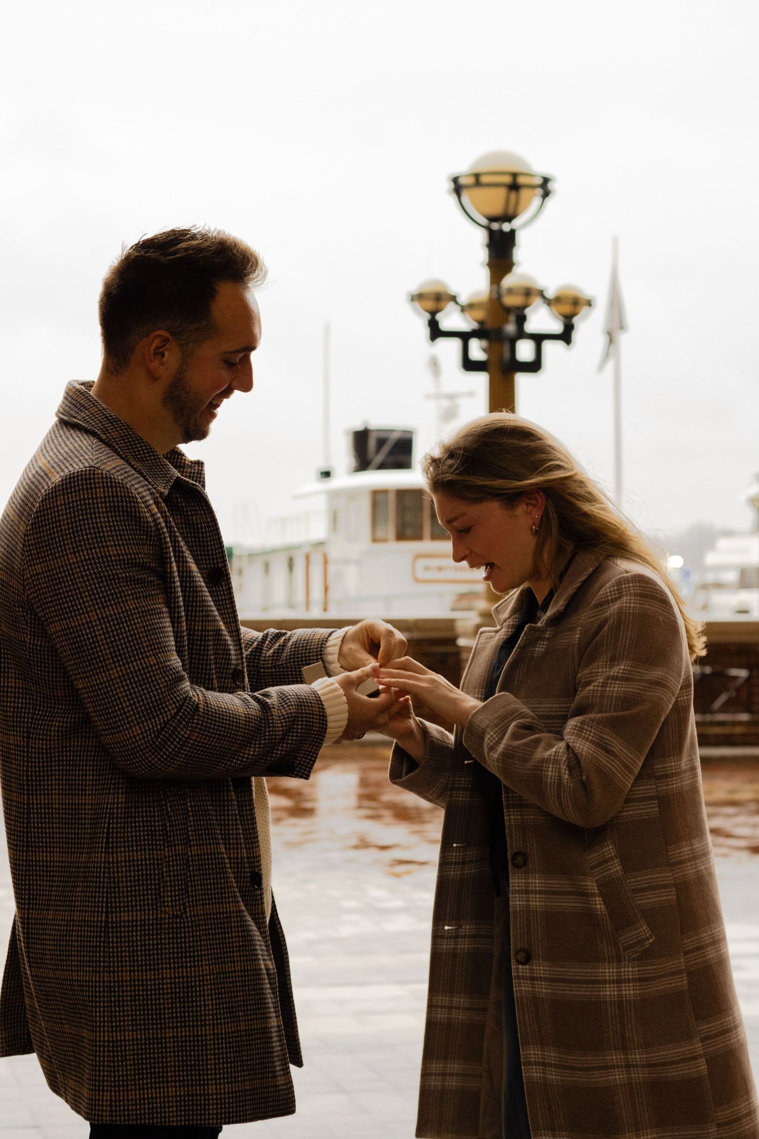 Man placing engagement ring on womans finger at rainy waterfront dock, both smiling joyfully.