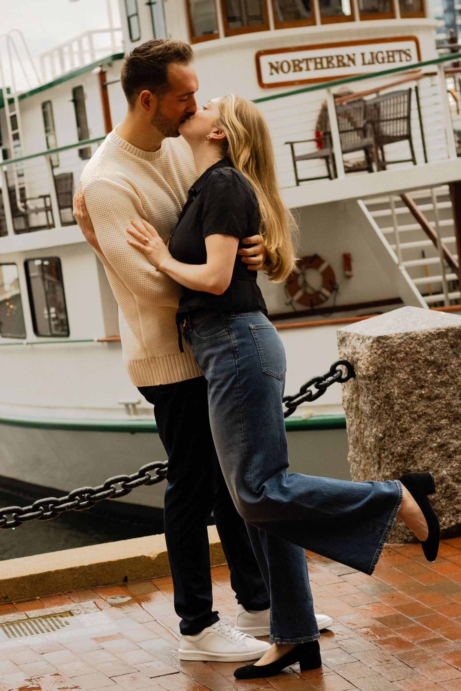 Couple sharing a tender kiss in front of the Northern Lights boat at a rainy harbor dock.