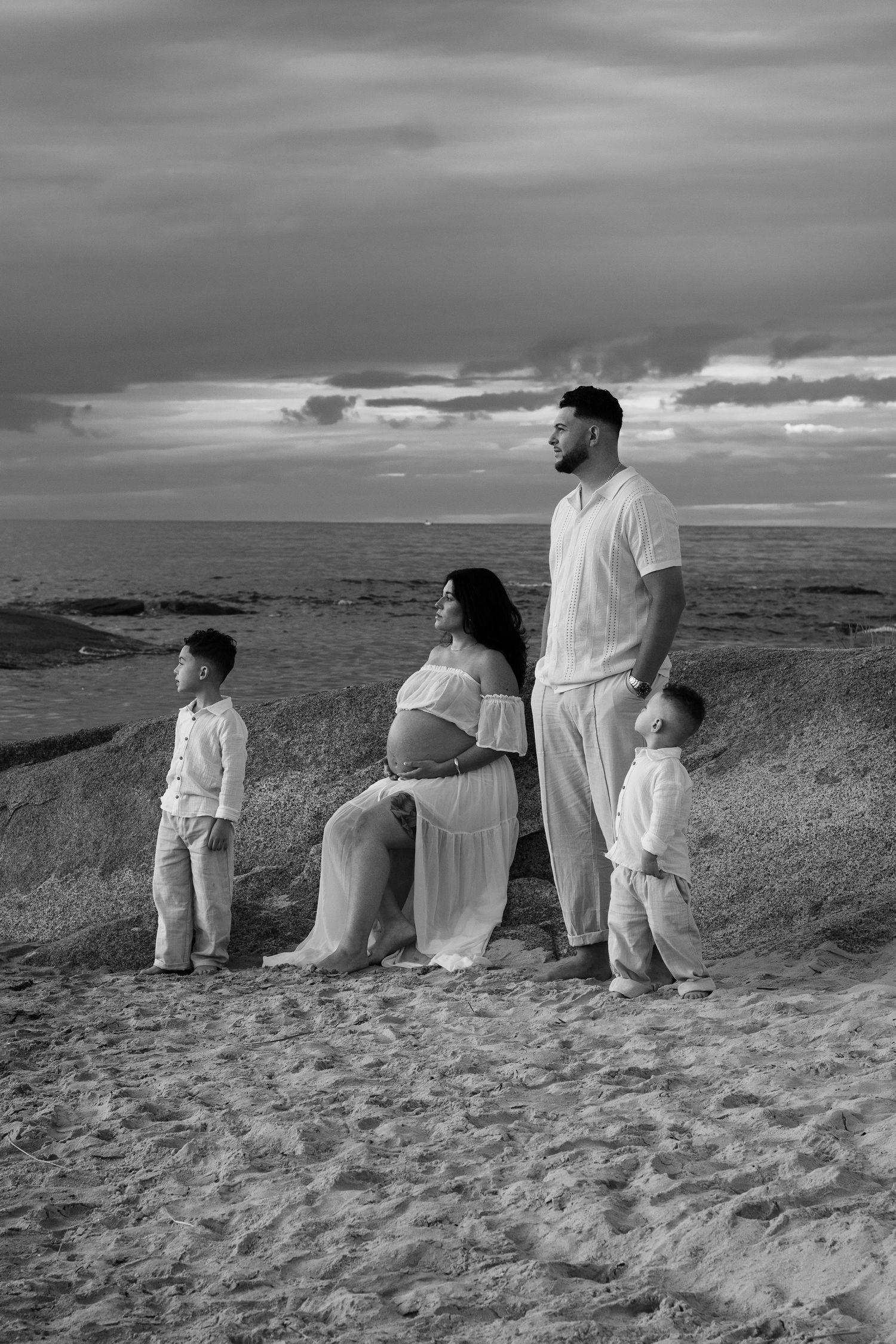 Black and white maternity photo of a family of four on a sandy beach at sunset, expecting mother in flowing gown.