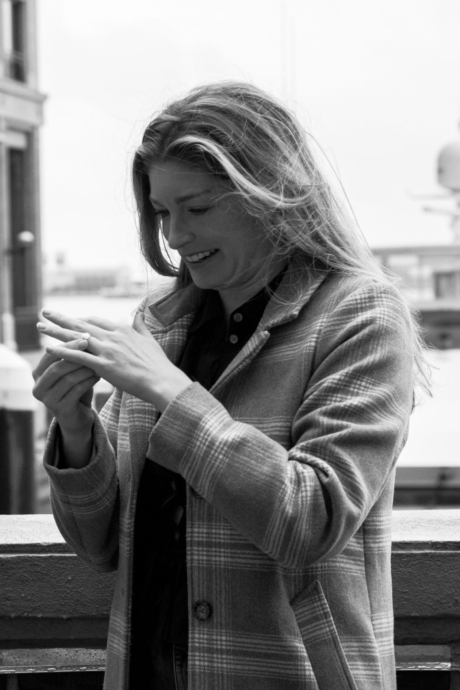 A black and white portrait of a blonde woman in a plaid coat looking at her ring outdoors.