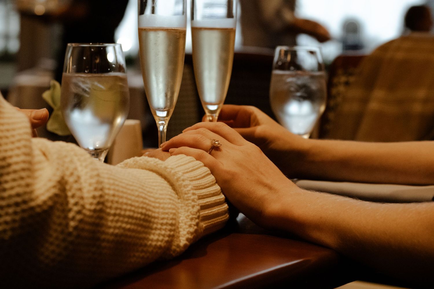 Close-up of an engaged couple holding hands beside champagne flutes at a restaurant table.
