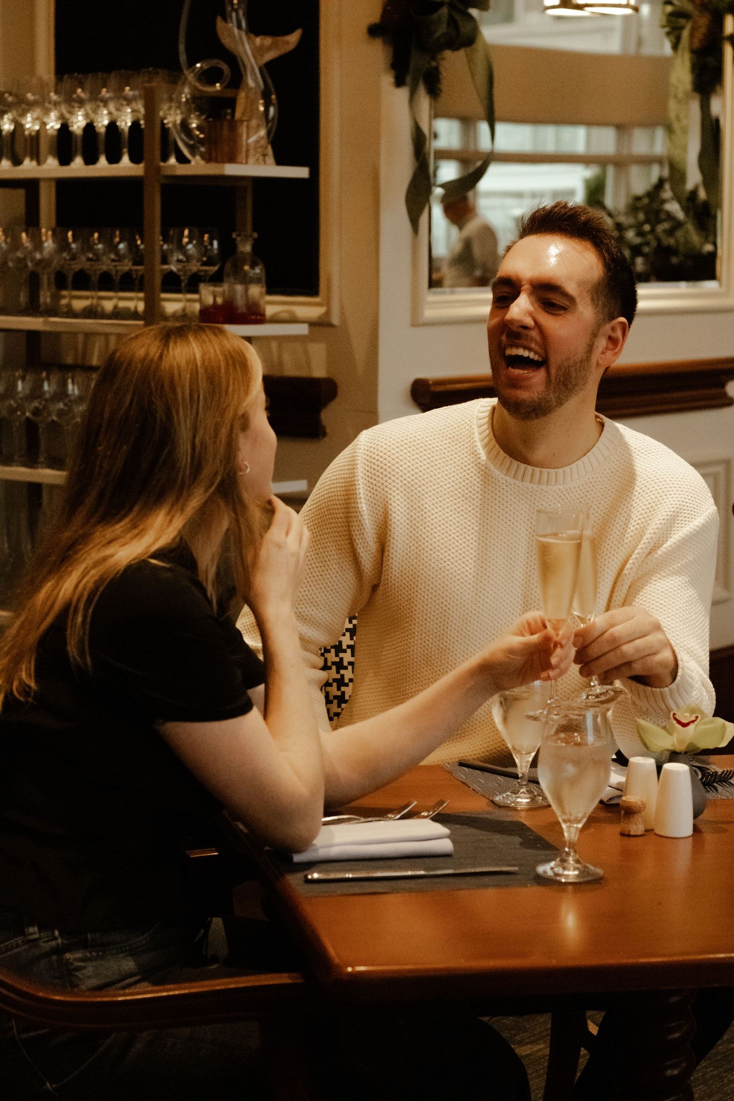 Joyful couple toasting with champagne glasses at a restaurant table, man laughing with delight.