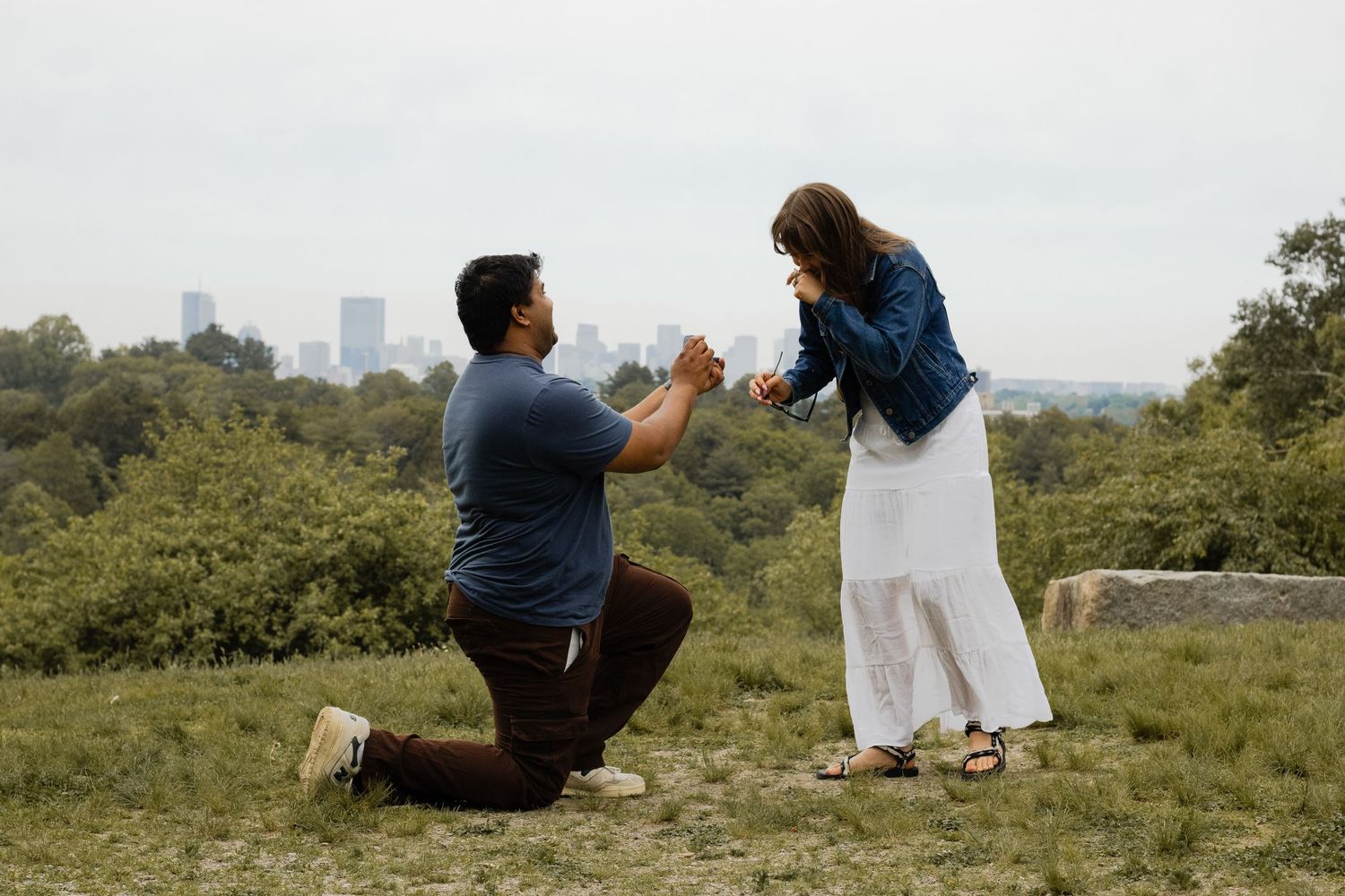 Man kneeling on one knee proposing to woman outdoors with city skyline in background on cloudy day.