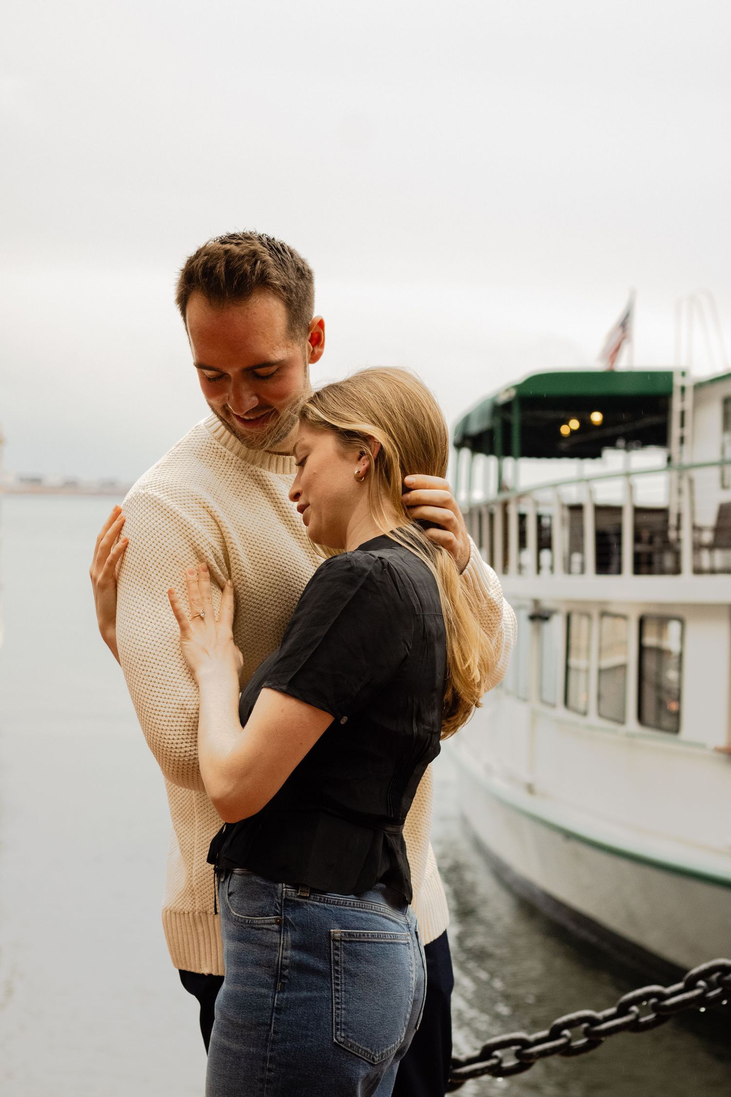 A couple embraces and smiles at each other near docked boats on a cloudy waterfront day.