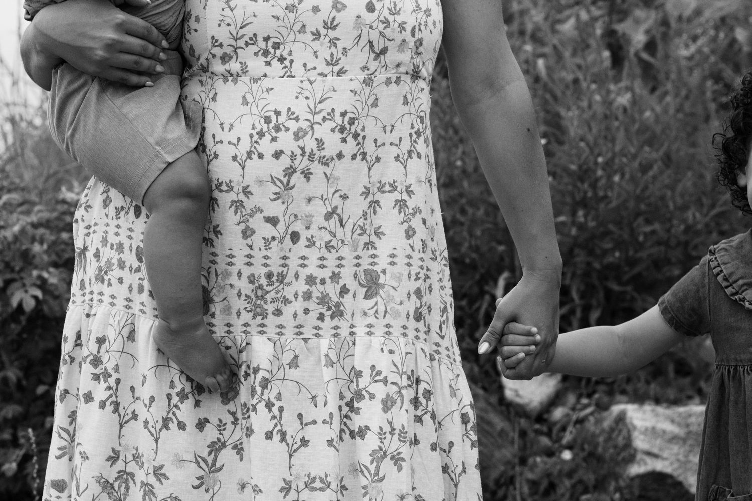 Black and white photo of a woman in floral dress holding a baby and a child's hand outdoors.
