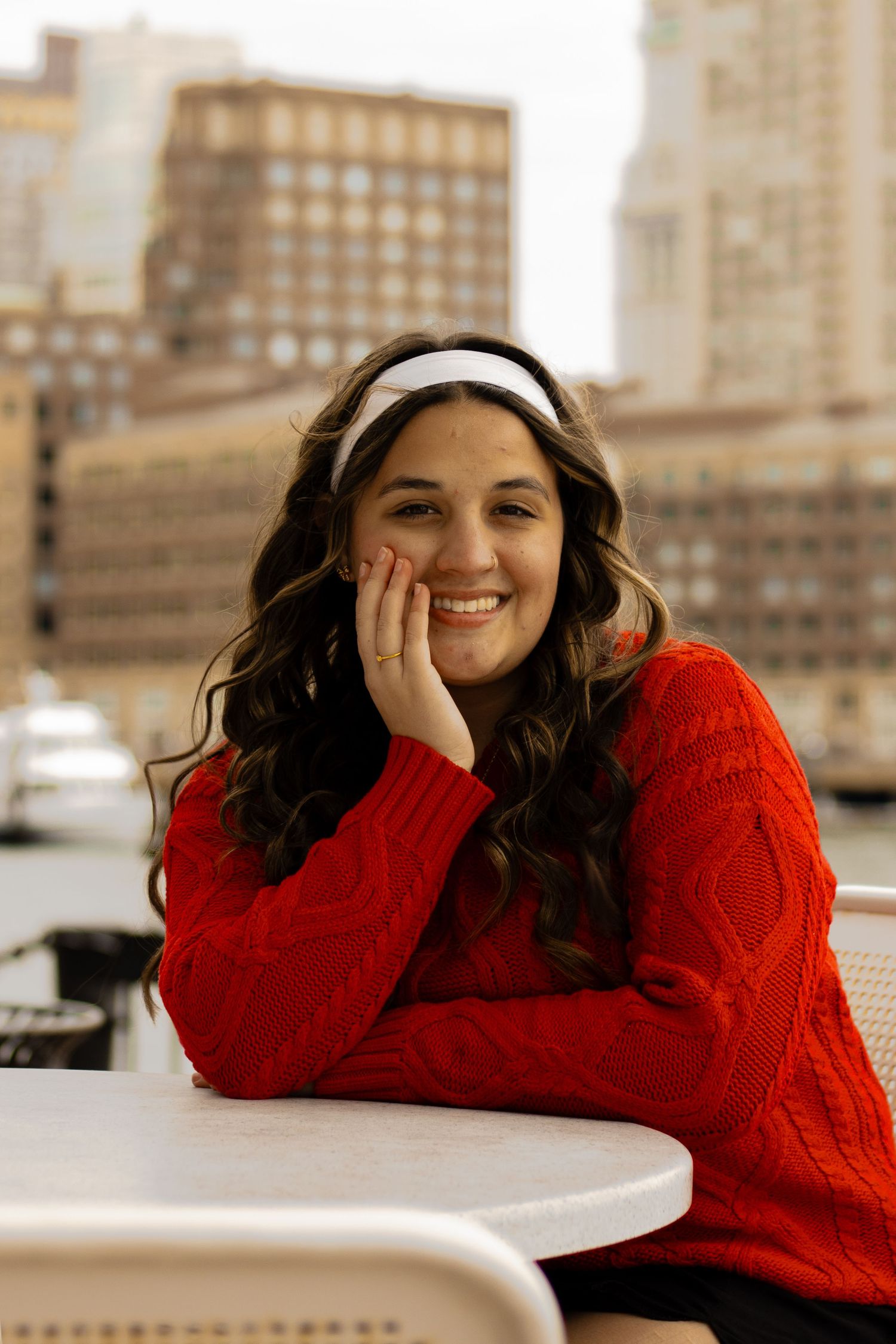 Smiling young woman in red cable-knit sweater and white headband leaning on outdoor table with city skyline backdrop.