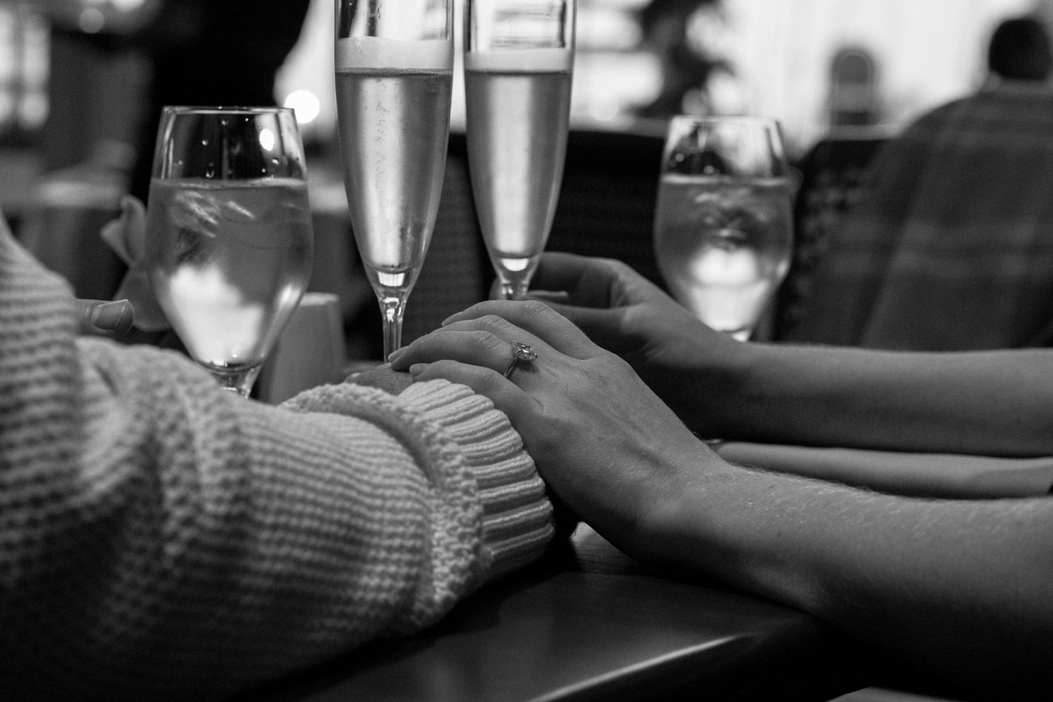 Close-up of couple holding hands at restaurant table with champagne flutes, engagement ring visible.