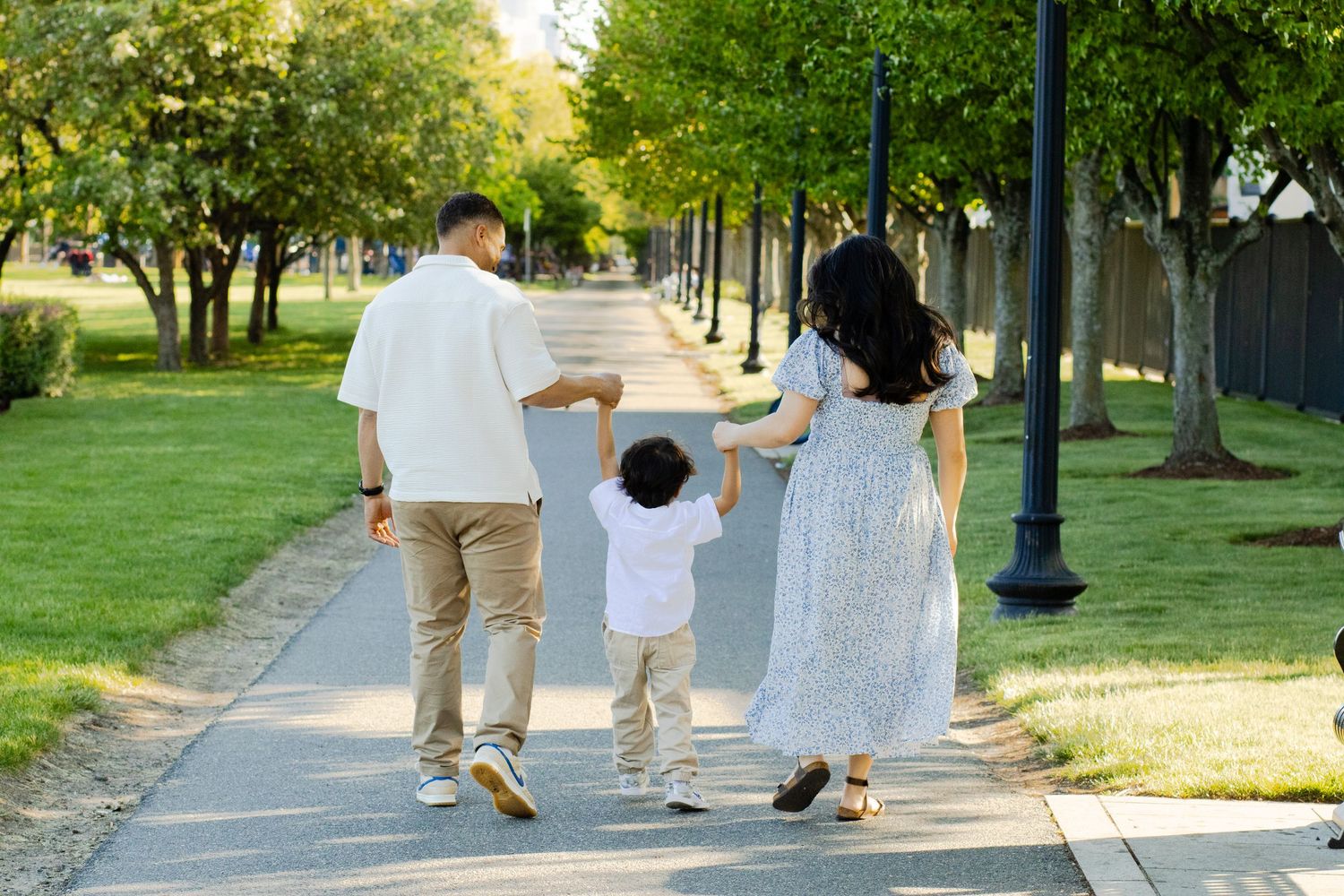 Family of three walking hand-in-hand down a tree-lined park path on a sunny day.