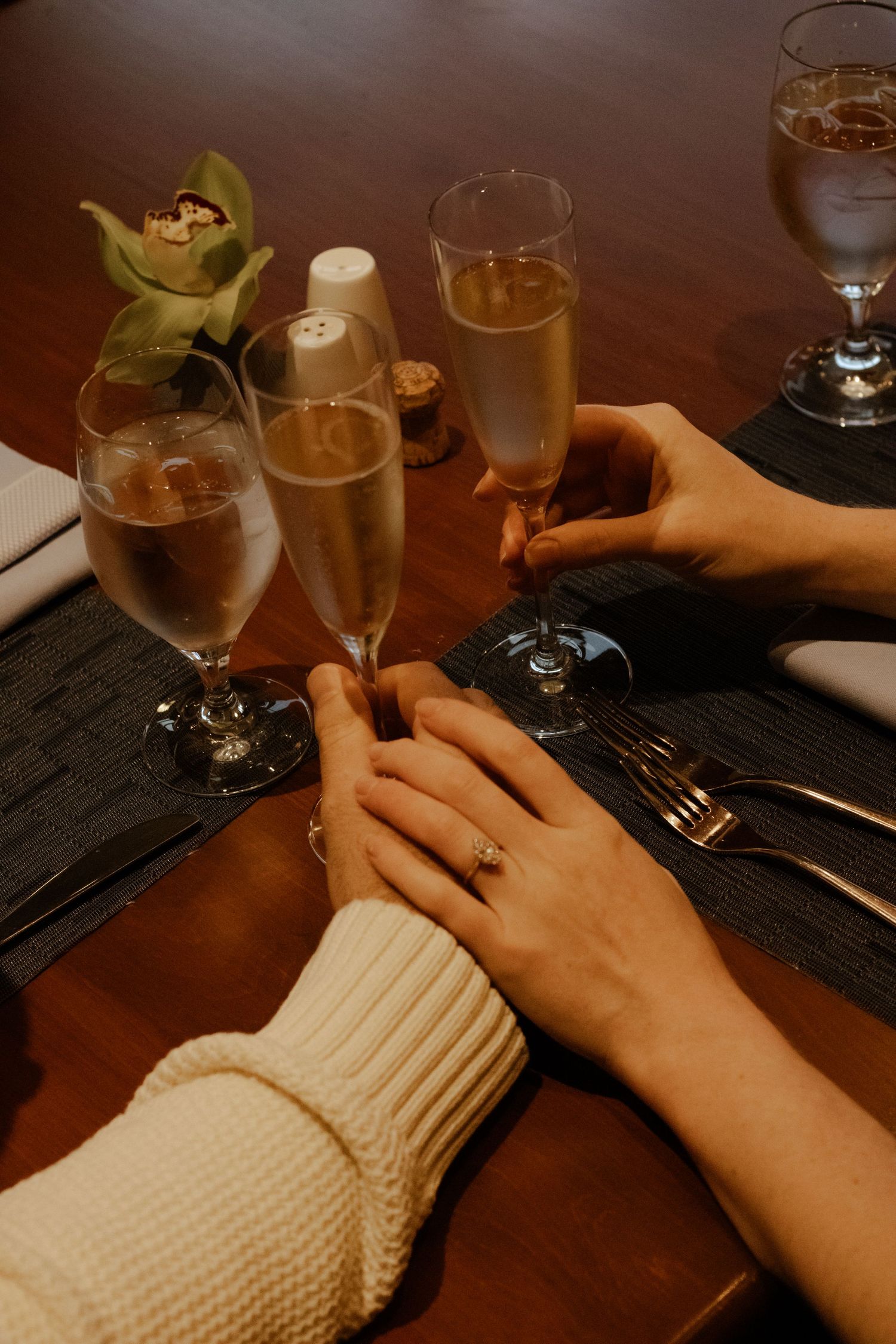 Engaged couple holding hands at a romantic dinner table with champagne flutes and an orchid centerpiece.