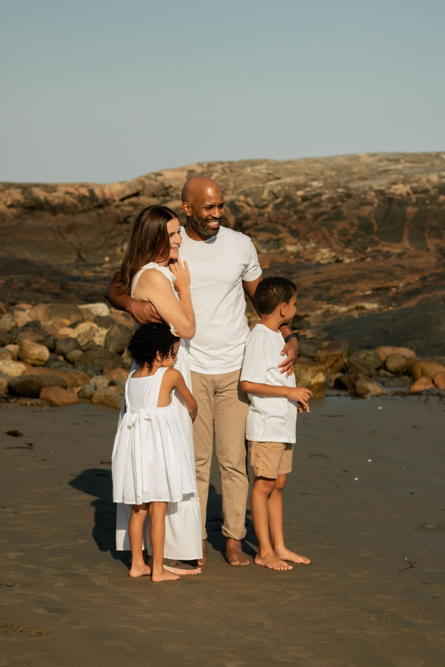 Happy family of four standing together on a beach at golden hour with rocky cliffs behind.