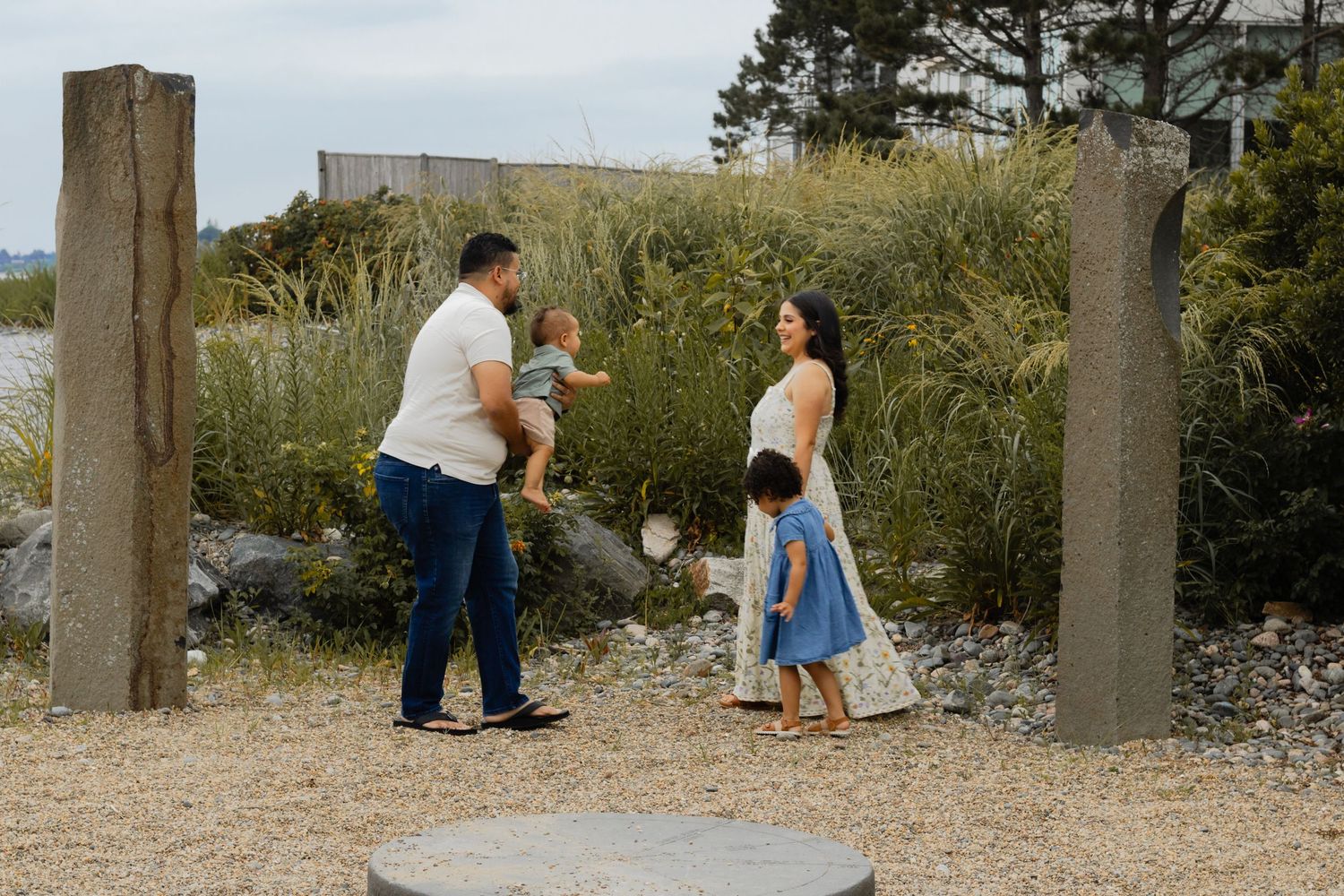 Family with two young children interacting outdoors near tall grasses and stone pillars.