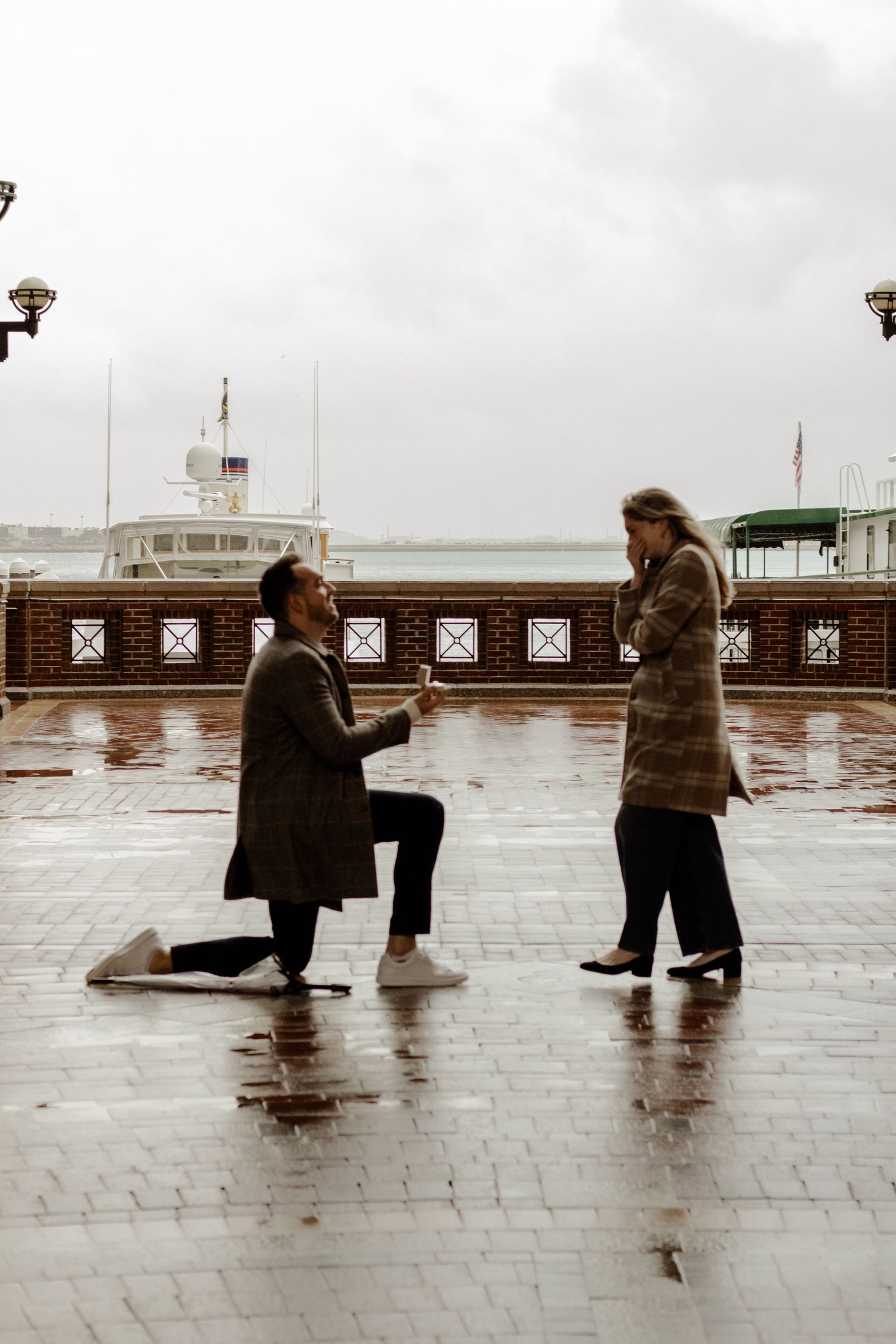 Romantic marriage proposal on wet pier, man kneeling with ring as woman reacts emotionally.