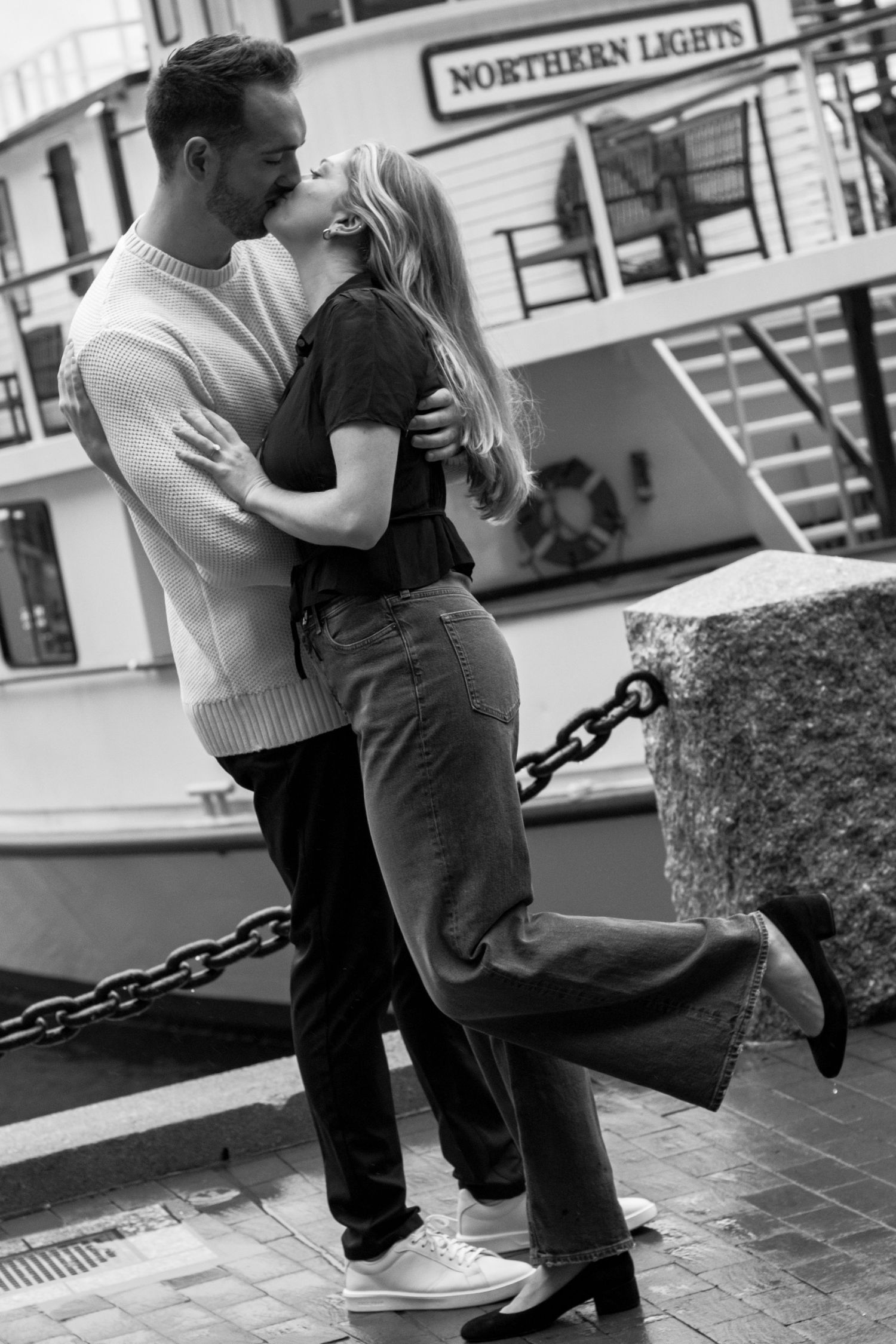 Black and white photo of a couple sharing a romantic kiss at a harbor dock with the Northern Lights boat behind them.
