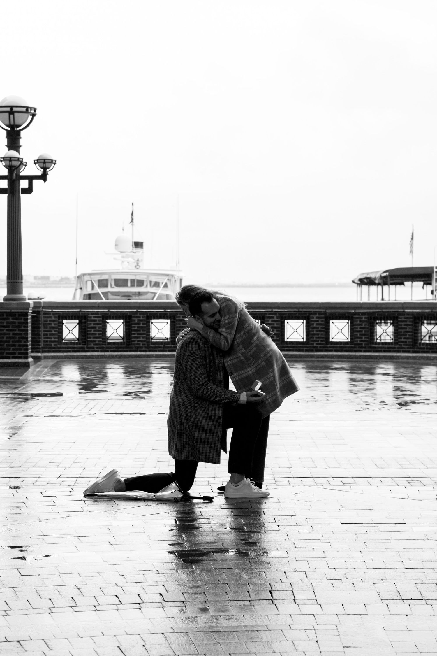 Black and white photo of couple embracing after marriage proposal on rainy pier.