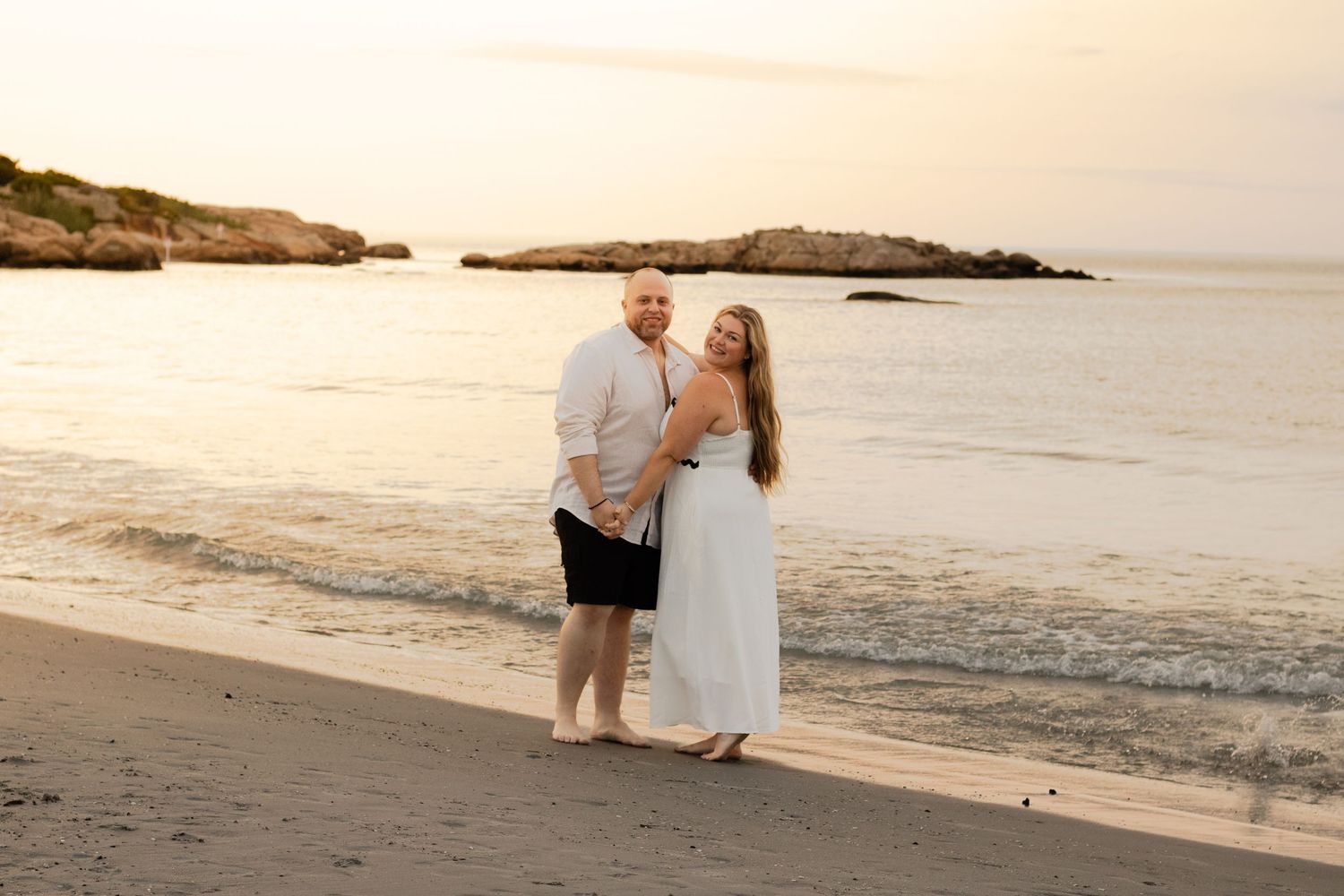 Couple embracing on sandy beach at sunset with rocky coastline in the background.