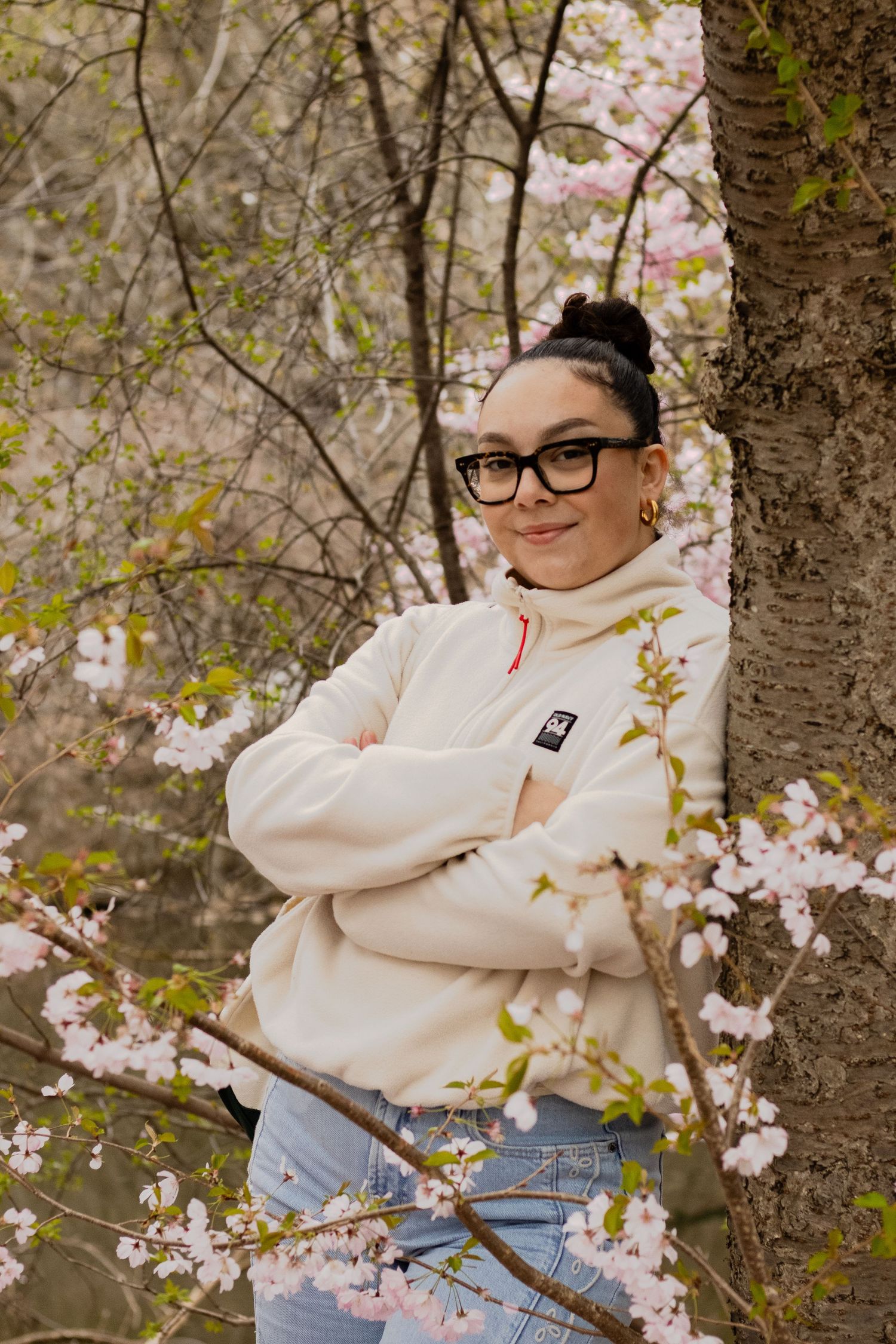 Young woman with glasses leaning against tree surrounded by pink cherry blossoms in spring.