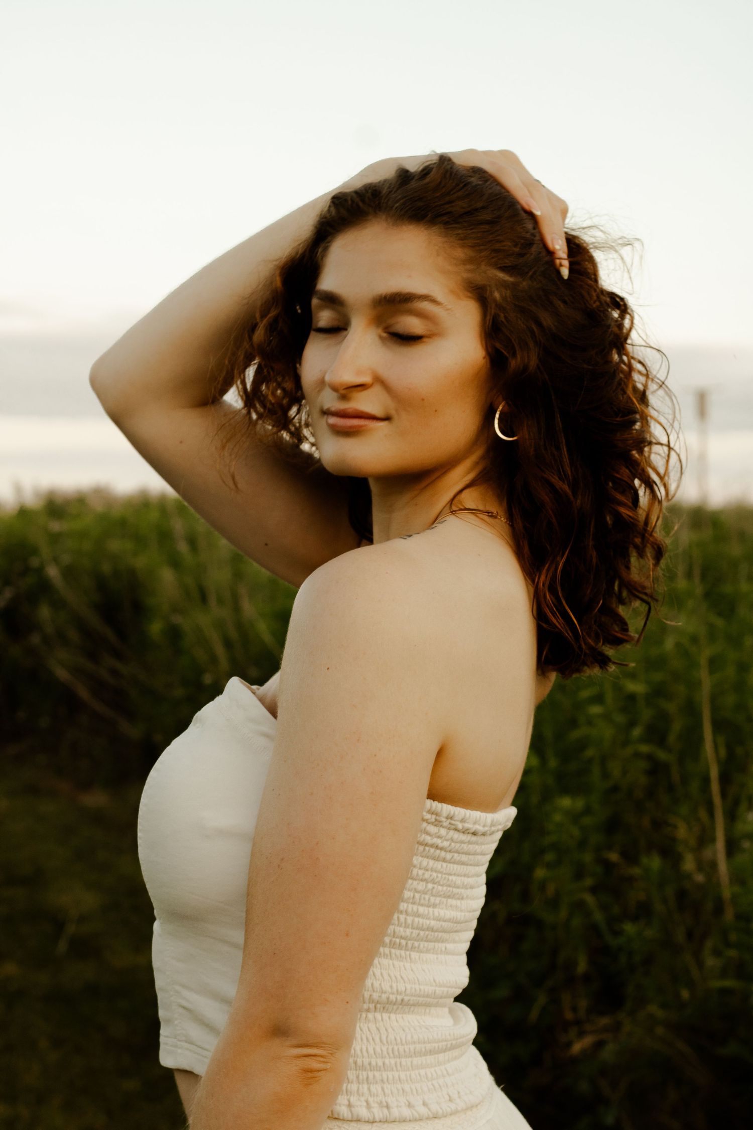 Young woman with curly hair in white smocked top poses outdoors at golden hour near green grass.