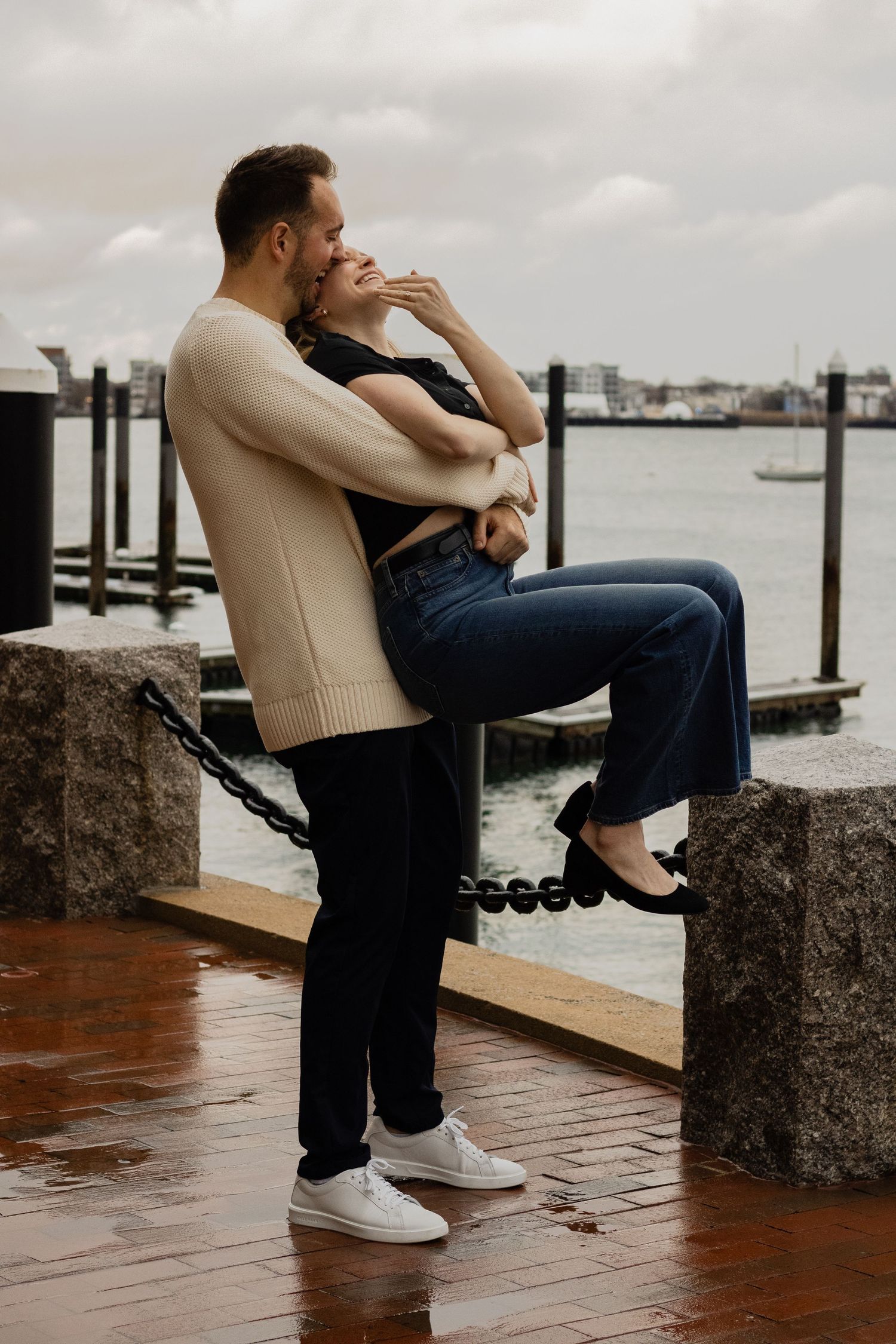 Man lifting his partner onto a harbor dock railing as they embrace on a rainy day by the water.