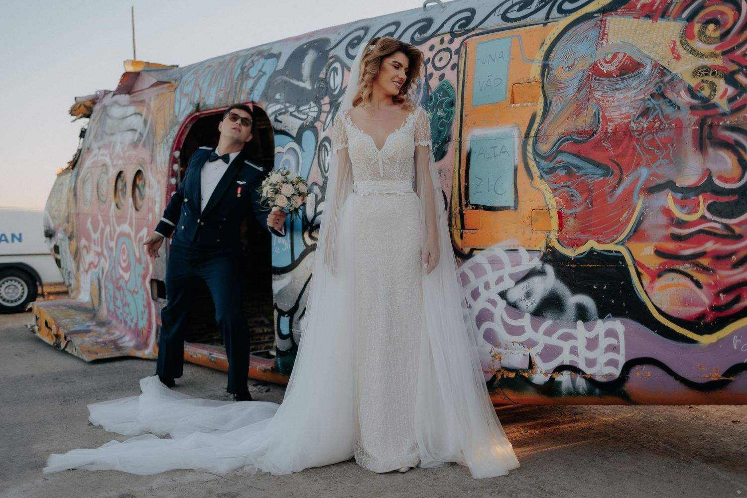Mireasă stând elegant în fața unui fuselaj cu graffiti, în timp ce mirele apare jucăuș din ușa avionului; moment creativ și vesel pe aerodro