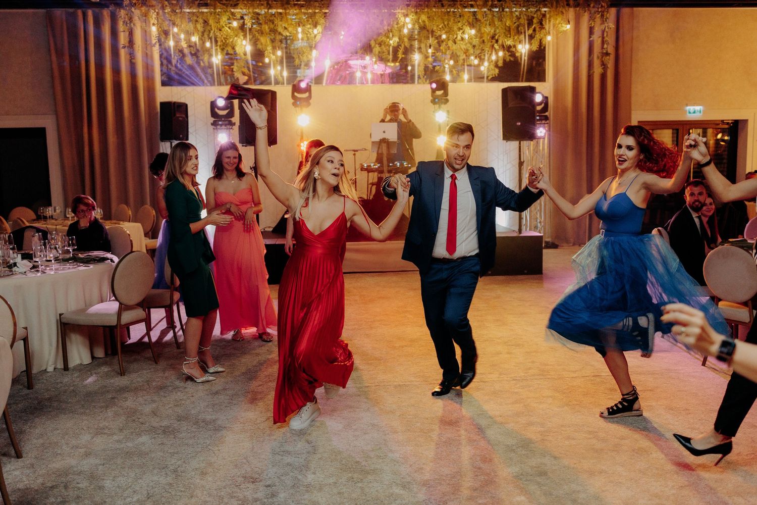 Grup de invitați dansând o horă plină de energie; atmosferă festivă și culori vibrante pe ringul de dans la o petrecere de nuntă.