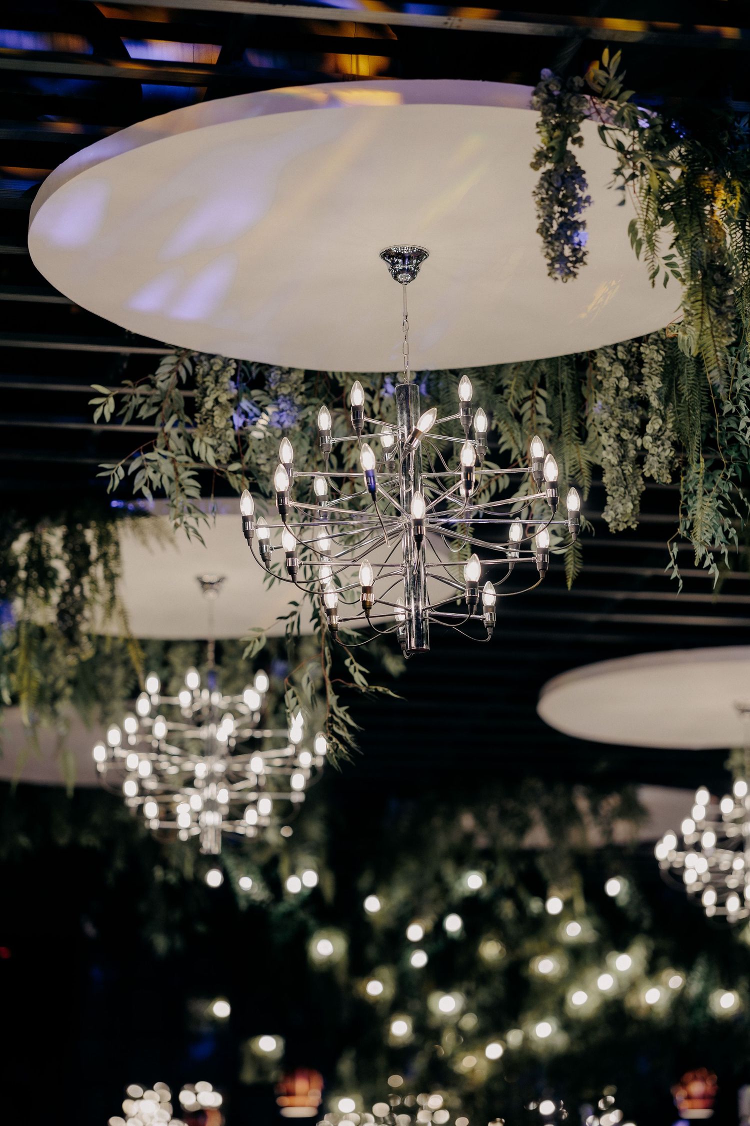 Candelabre moderne și elegante suspendate printre aranjamente de verdeață naturală, creând un decor sofisticat în sala de nuntă.