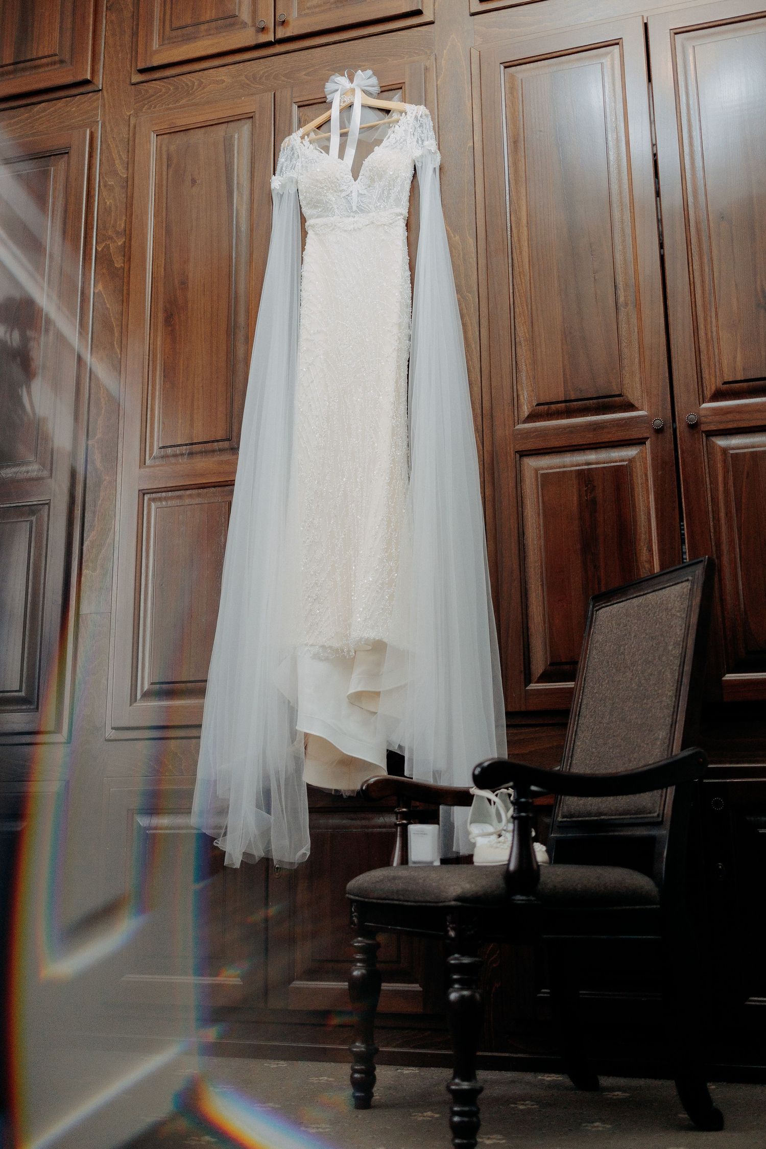Rochie de mireasă albă cu mâneci lungi din tull, suspendată pe un dulap masiv din lemn; detaliu elegant înaintea pregătirilor de nuntă.