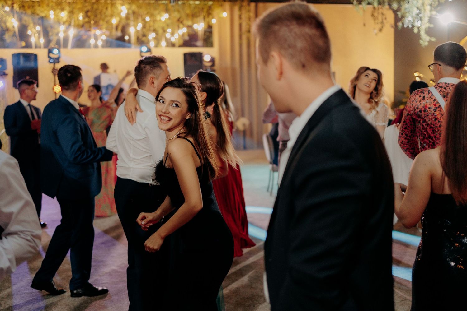 Invitată zâmbitoare în rochie neagră dansând pe ringul de dans; privire veselă spre cameră într-o atmosferă festivă de nuntă.