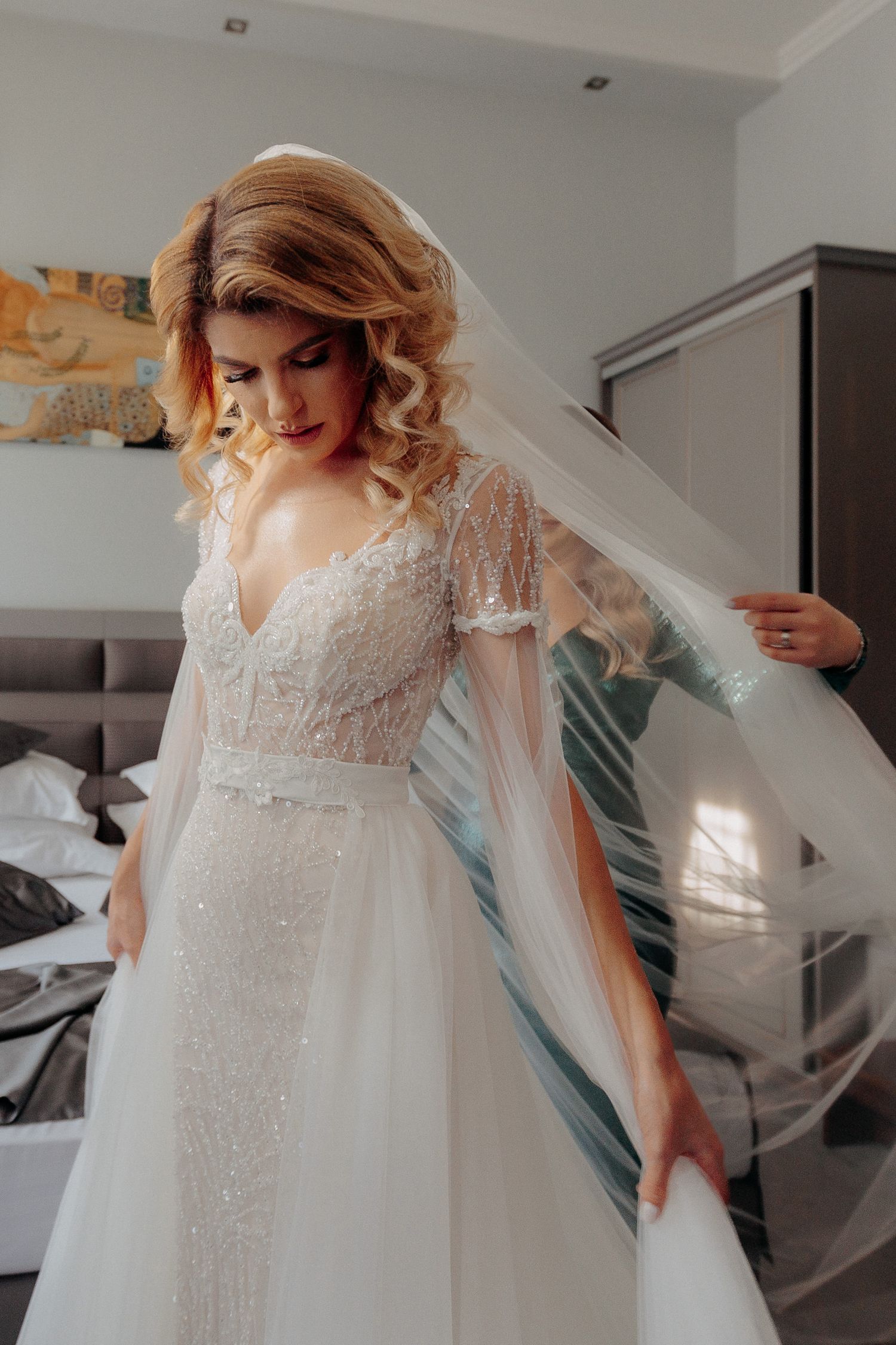 Portret cu mireasa în rochie albă strălucitoare, având vălul lung aranjat de o domnișoară de onoare în timpul pregătirilor de nuntă.