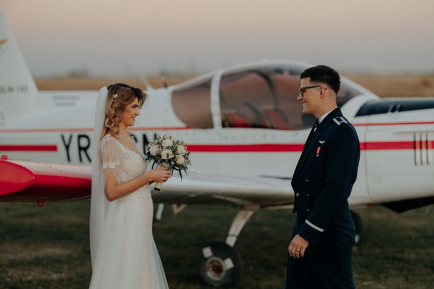 Miri zâmbindu-și complice lângă un avion pe aerodrom; mireasa poartă un buchet alb, iar mirele este în uniformă, într-un cadru romantic.