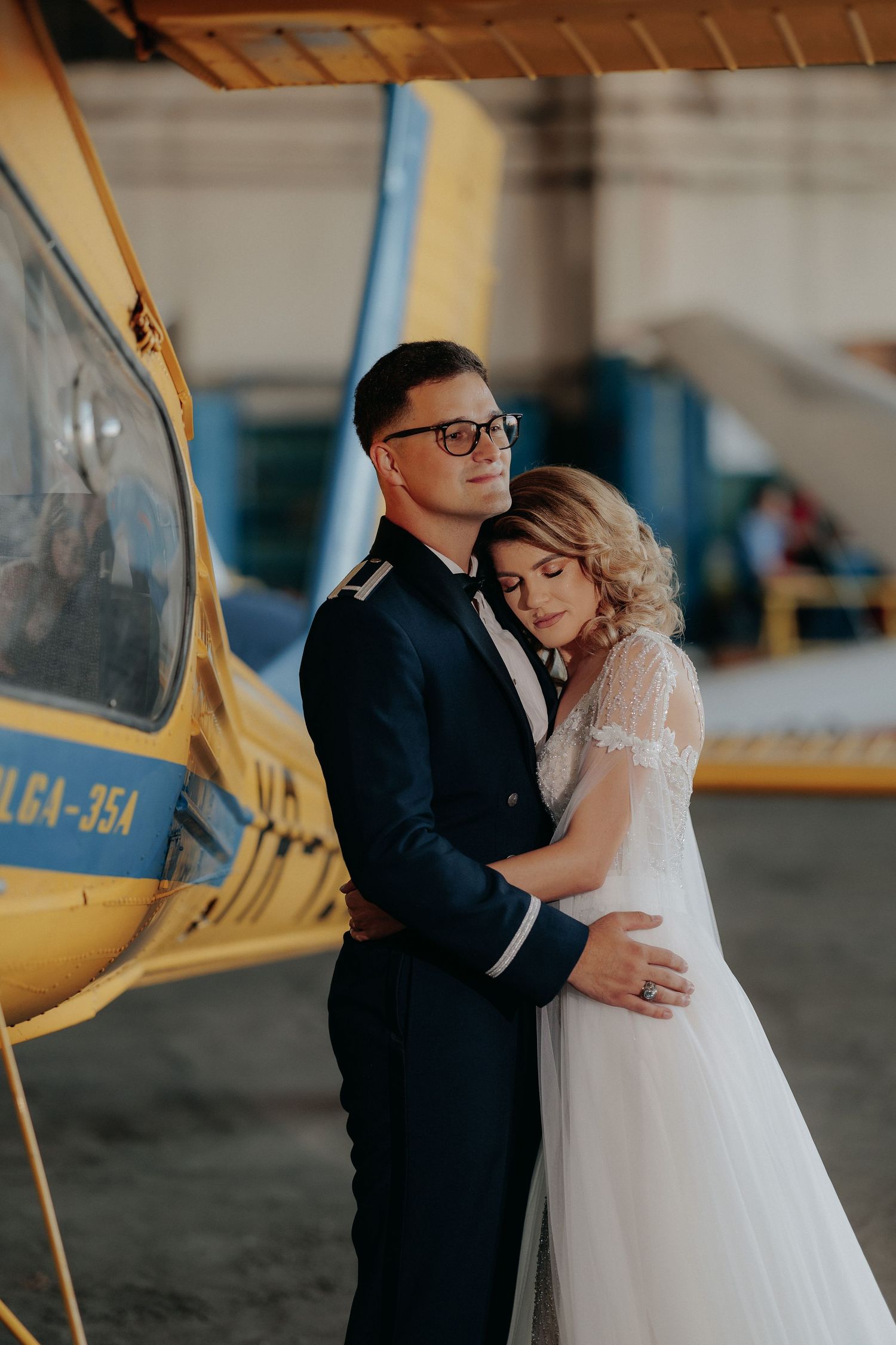 Mire îmbrățișându-și mireasa lângă un avion galben într-un hangar; mireasa își sprijină capul pe umărul lui, într-un moment de liniște și af
