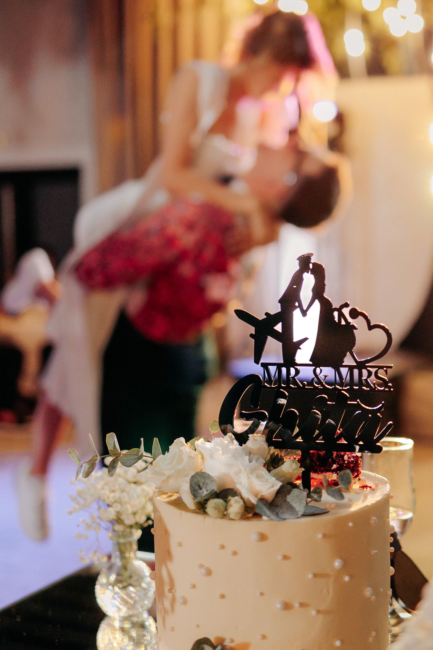 Tort de nuntă elegant cu topper „Mr. & Mrs. Ghiță”; mirii sărutându-se în fundal, într-un cadru de poveste cu lumini calde.