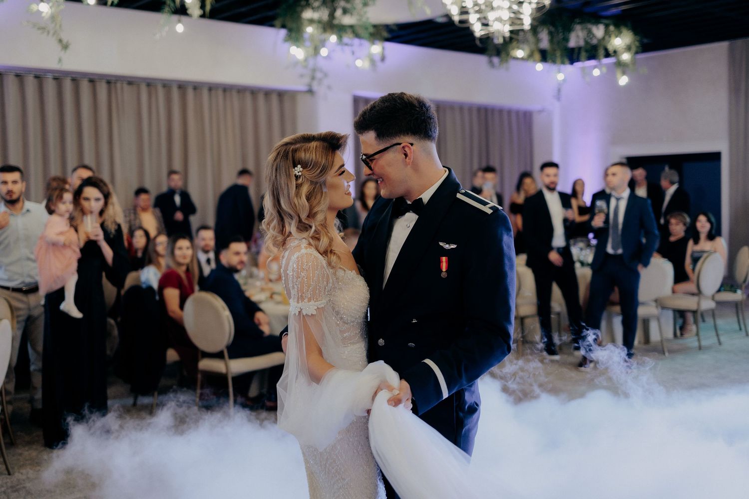 Miri privindu-se cu dragoste în timp ce dansează pe ringul acoperit de fum; moment romantic și plin de emoție la o nuntă elegantă.