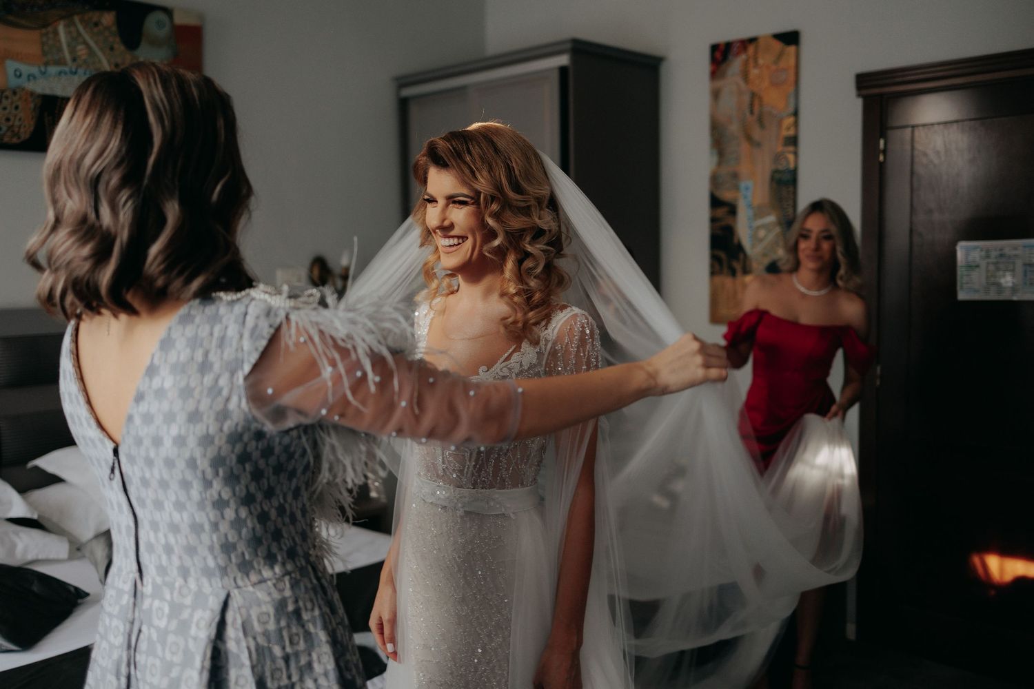Mireasă zâmbitoare ajutată de domnișoare de onoare să își aranjeze vălul lung; moment vesel din timpul pregătirilor de nuntă în dormitor.