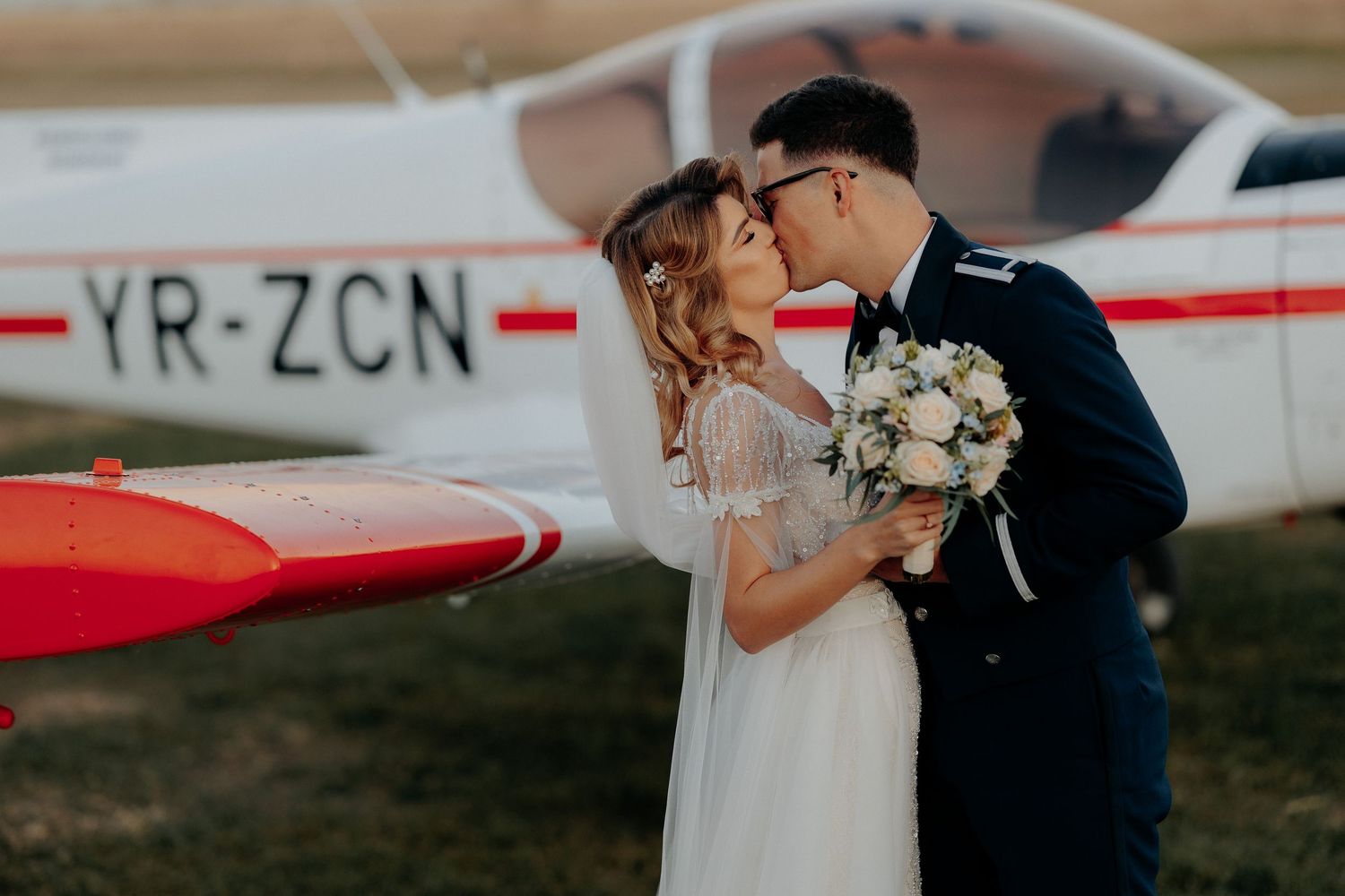 Miri sărutându-se romantic în fața unui avion alb de mici dimensiuni; mireasa ține un buchet de trandafiri, cadru elegant în aer liber.